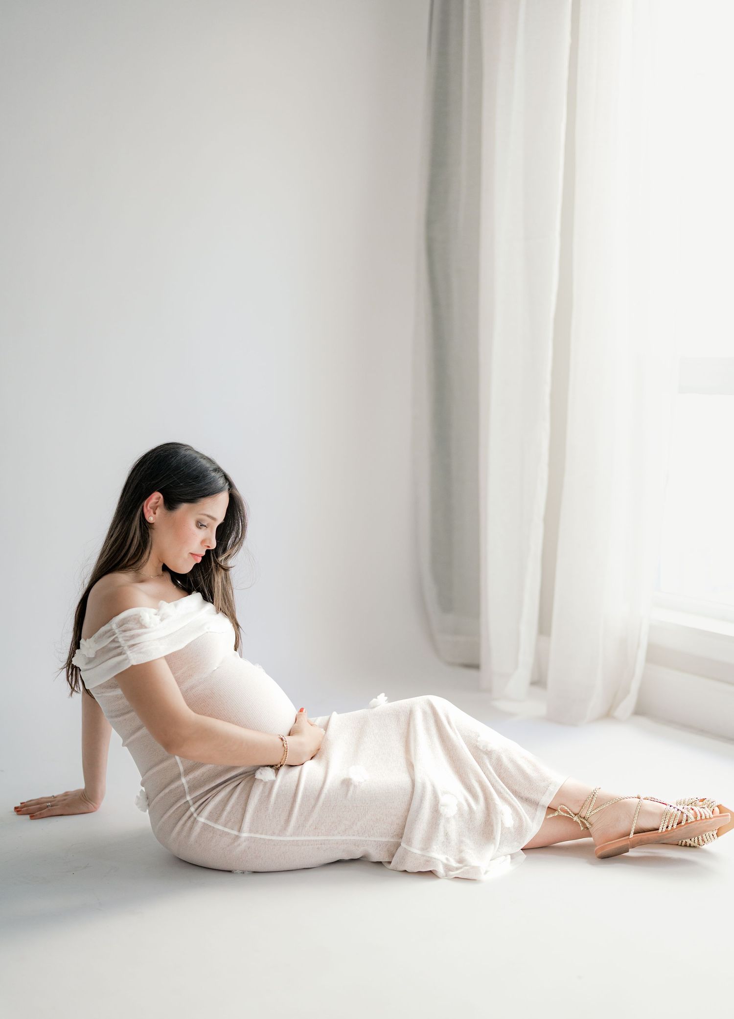 Artistic maternity photos of woman in white dress reclining against bright window light.