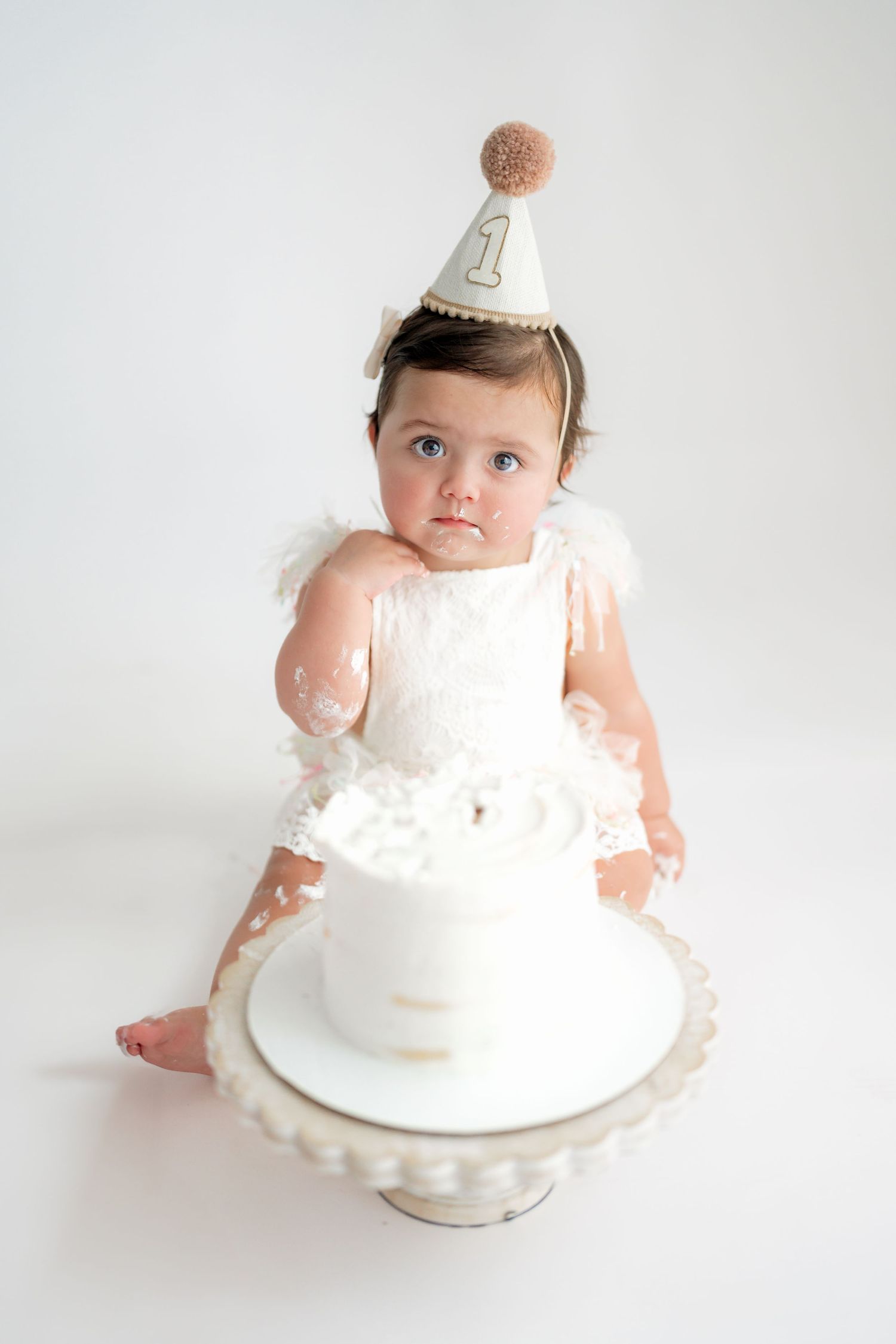 Baby in white dress and party hat sits surrounded by cake smash photos on white background.