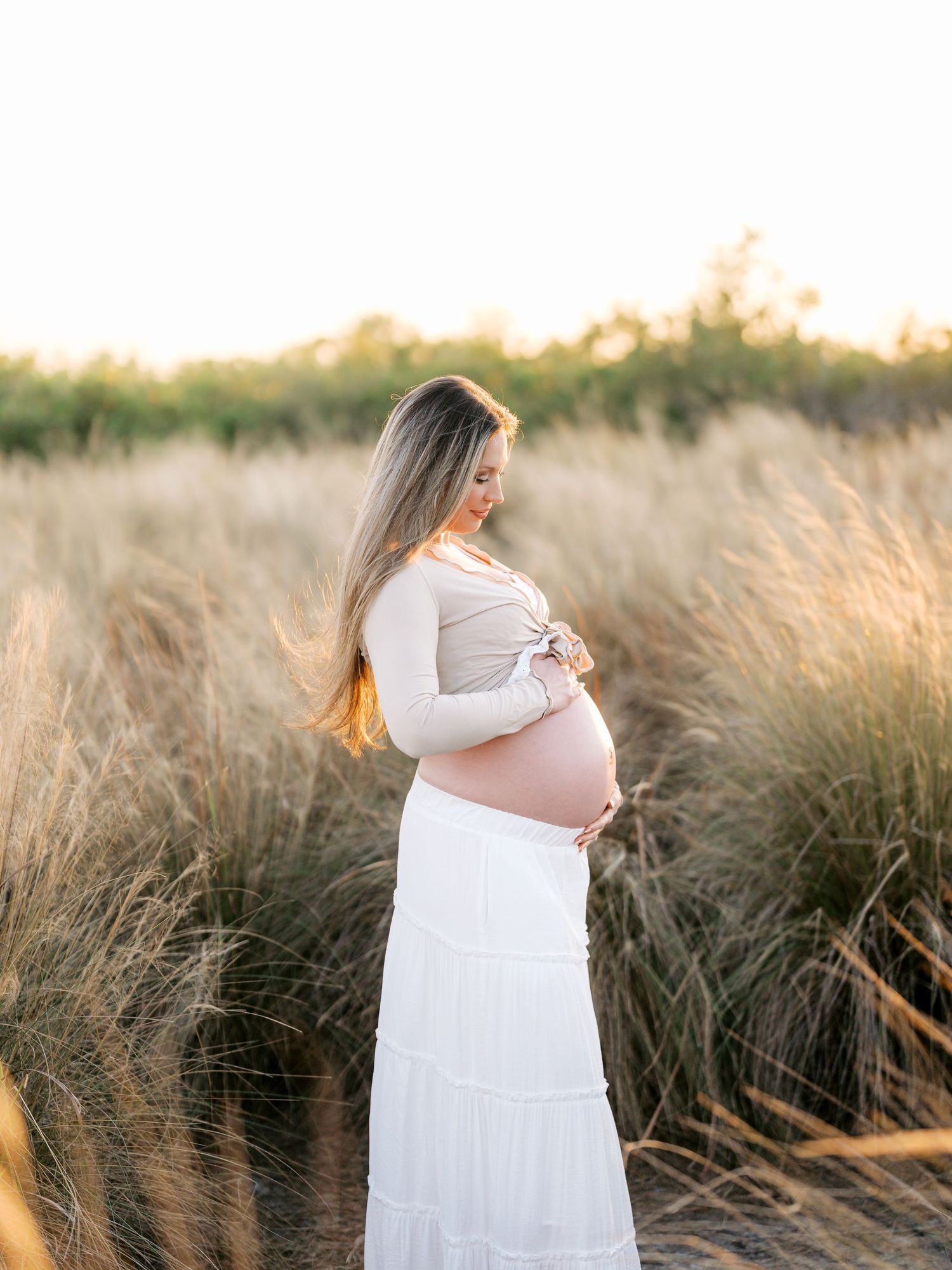 Pregnant woman in white skirt and beige top cradling baby bump in golden field at sunset.