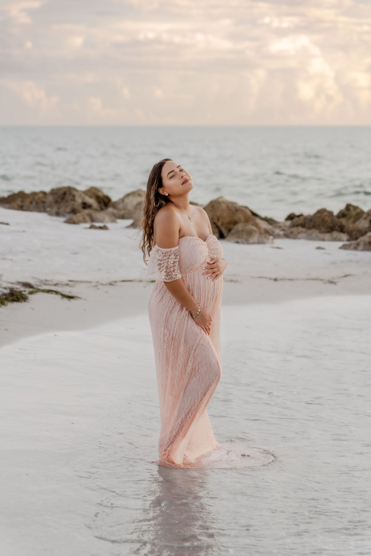 A figure in a flowing peach dress stands gracefully in shallow ocean waters at sunset along a rocky beach.