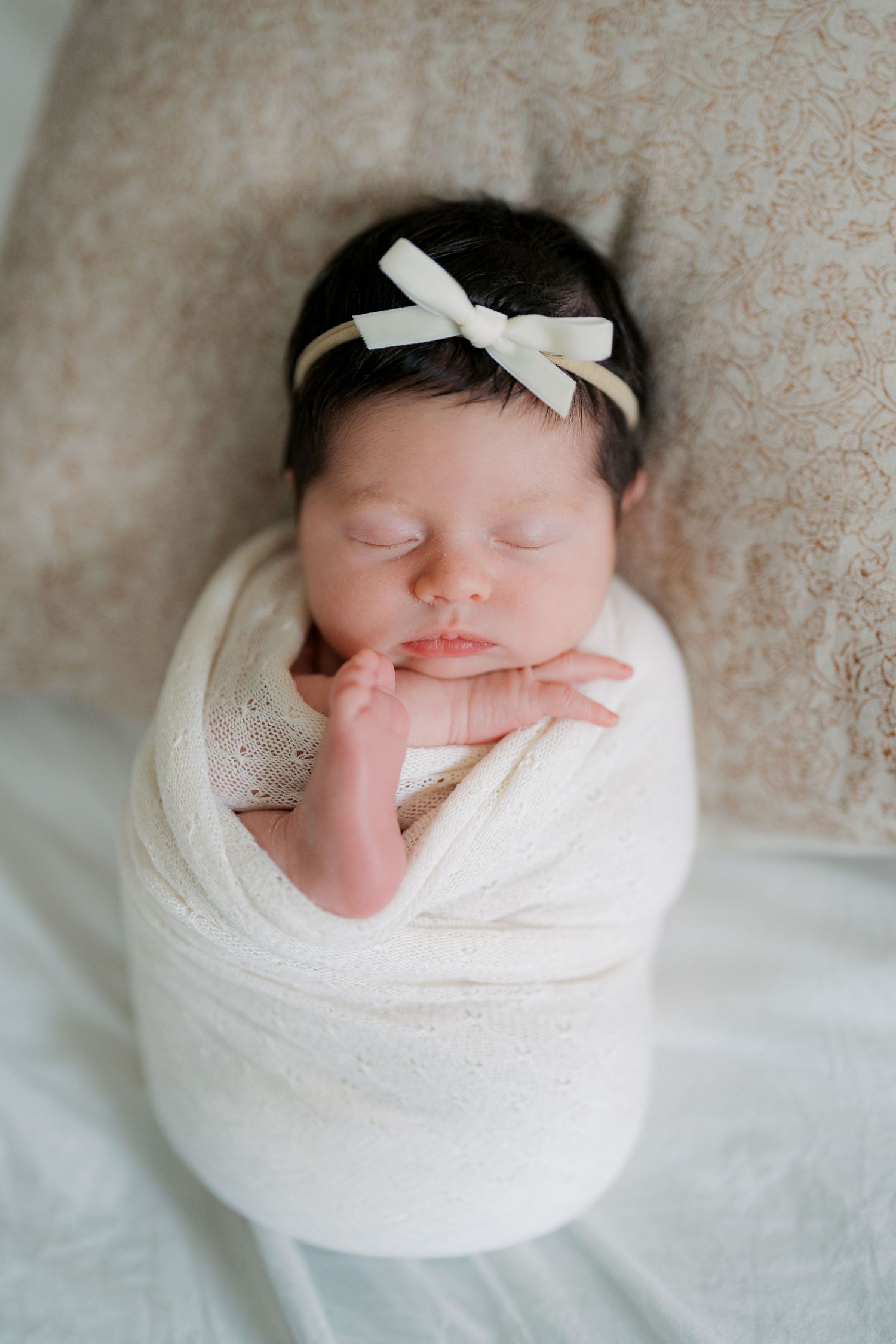 Sleeping newborn baby swaddled in white wrap with bow headband, peaceful infant portrait photography.