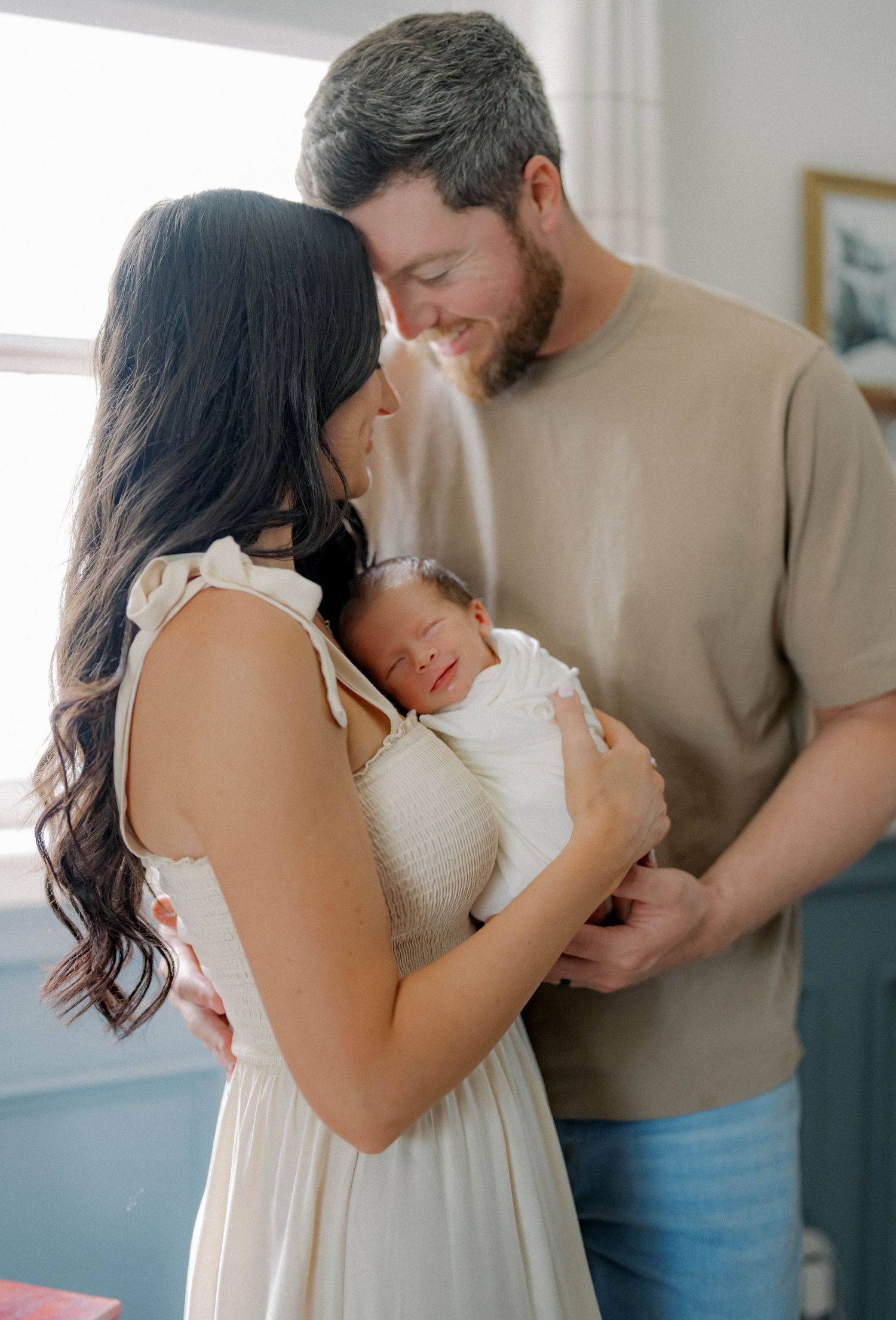 A couple tenderly holds their newborn baby, the woman in a white dress and man in a tan shirt, sharing an intimate family moment.