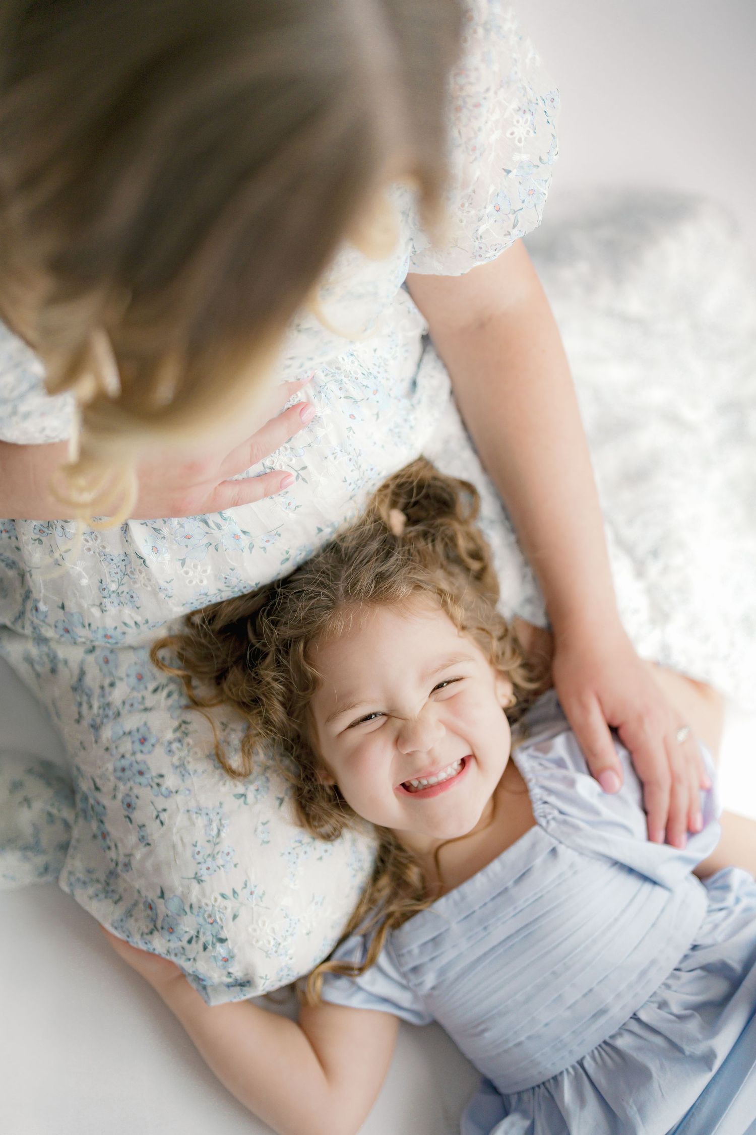 Little one lying down and smiling with a pregnant belly in frame wearing a blue floral dress.