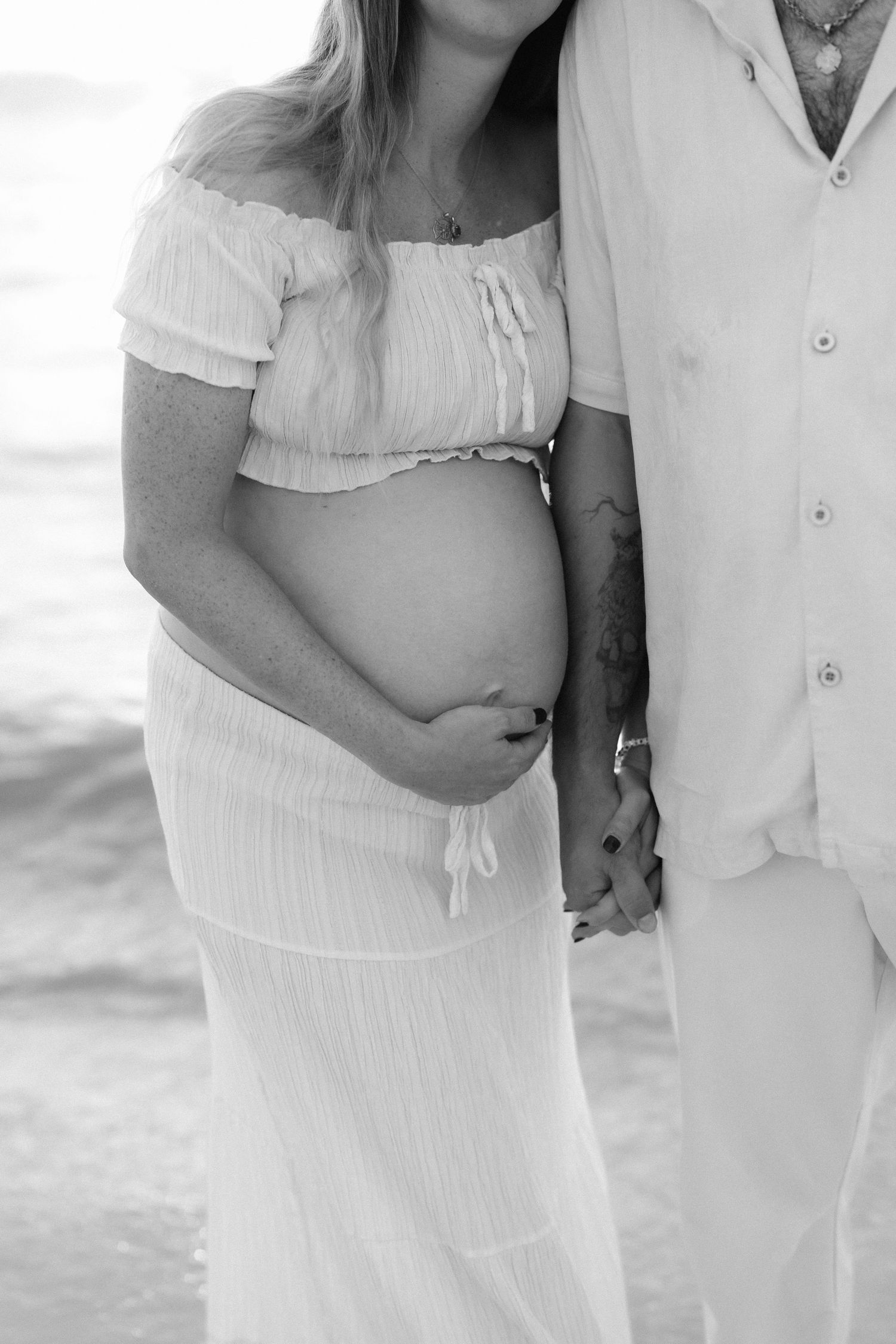 Black and white maternity photo of pregnant woman in white dress holding belly with partner at beach.