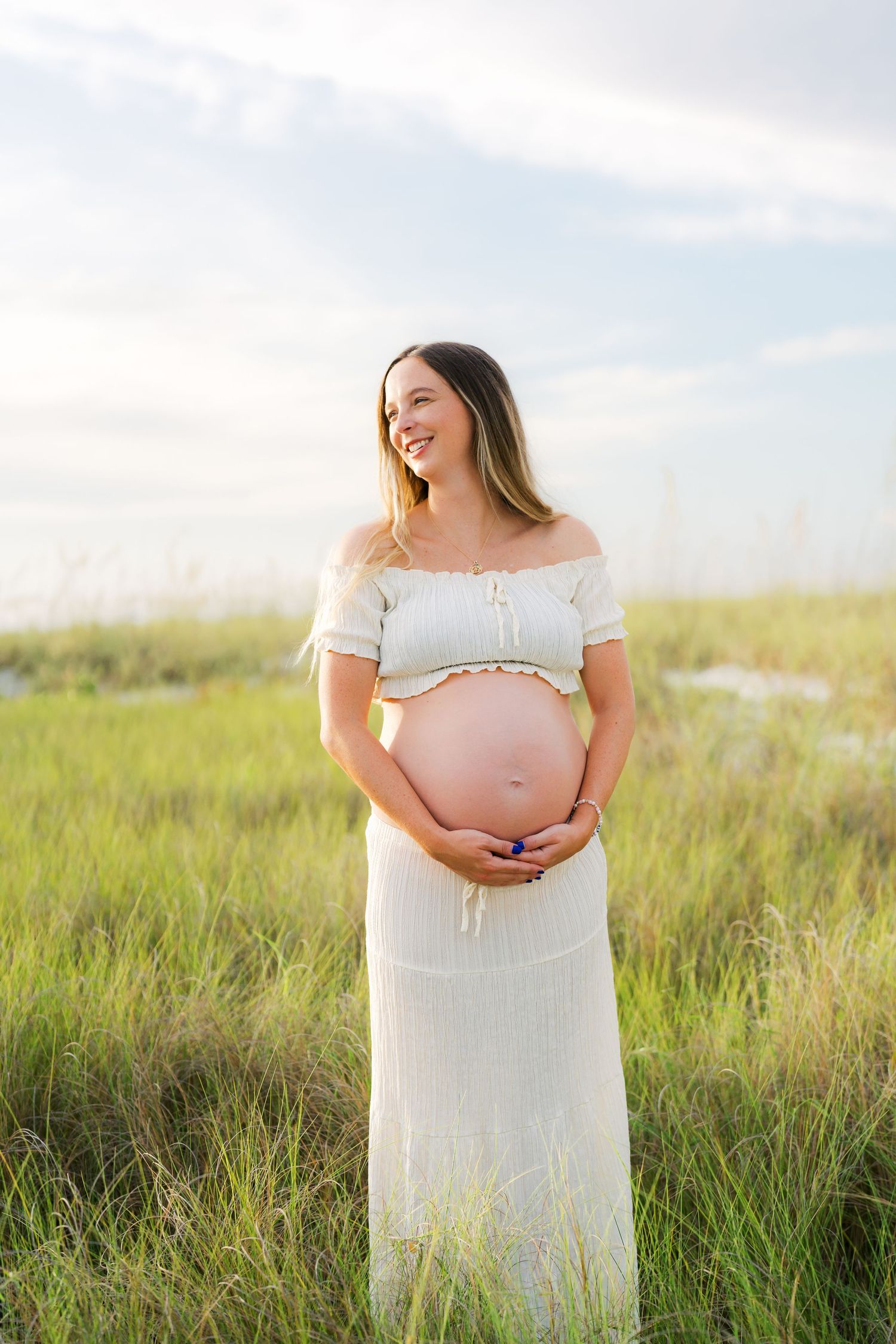 Smiling pregnant woman in white crop top and skirt cradling her baby bump in a sunlit green field.