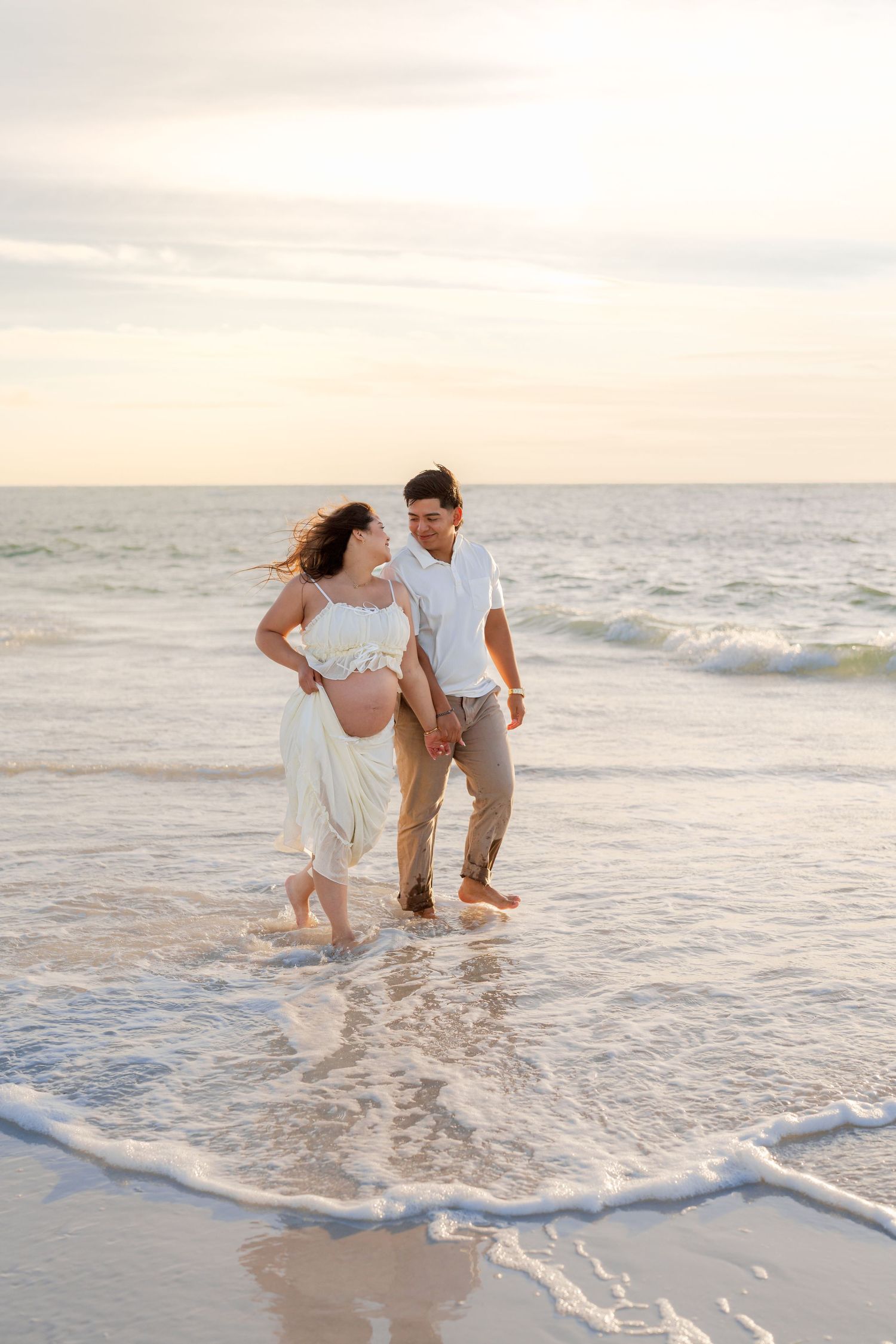 A couple walking and splashing in the ocean waves at sunset on a sandy beach during a romantic photoshoot.
