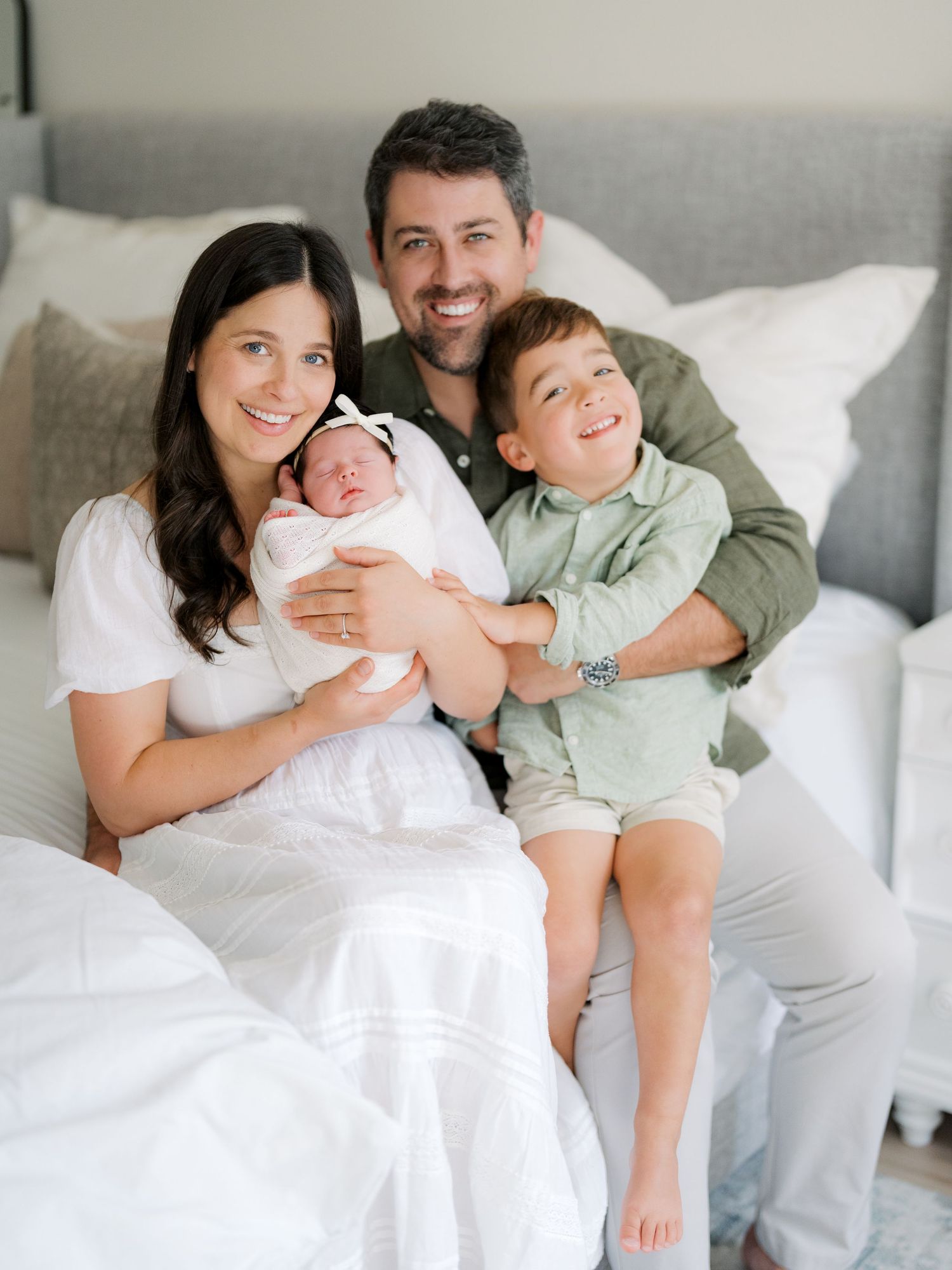 A happy family of four poses on a bed, with mom holding a newborn baby, dad, and young son smiling together.