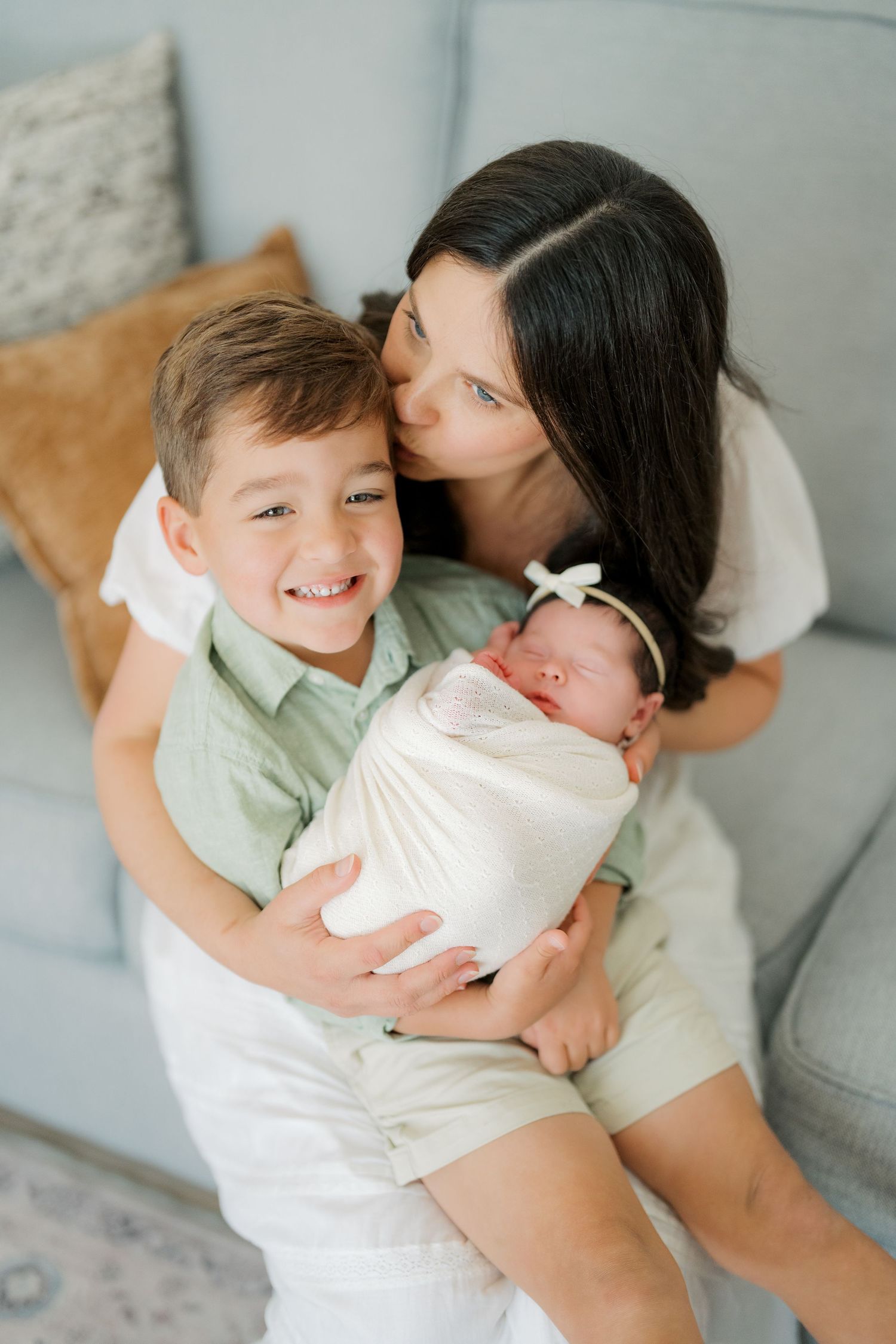 A mother kisses her smiling young son as he holds a swaddled newborn baby, sitting together by a light blue sofa.
