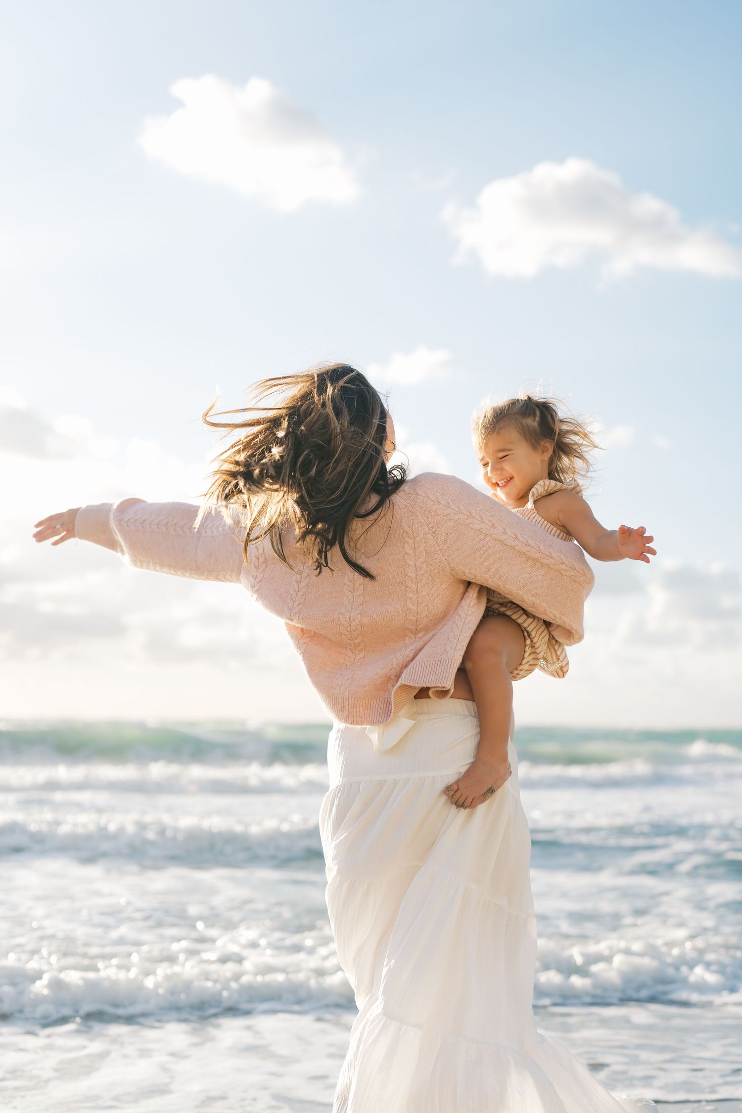 Mother holding young daughter on a sunny beach, both with arms outstretched, waves crashing behind them.