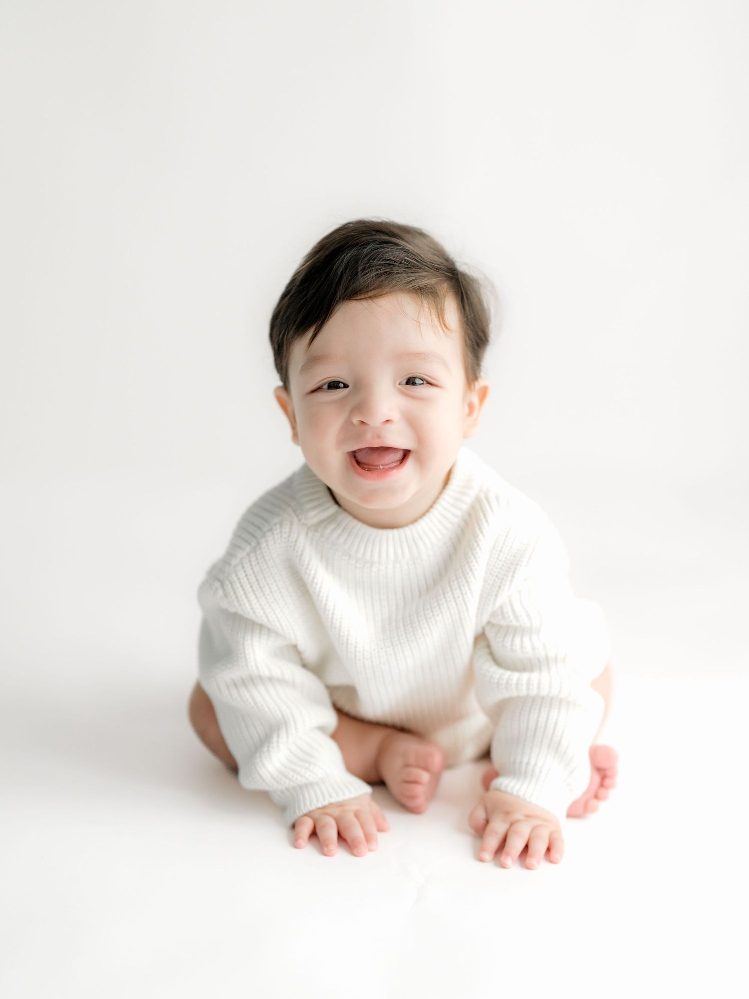 A smiling baby in a white knit sweater crawls and plays against a bright white studio backdrop.