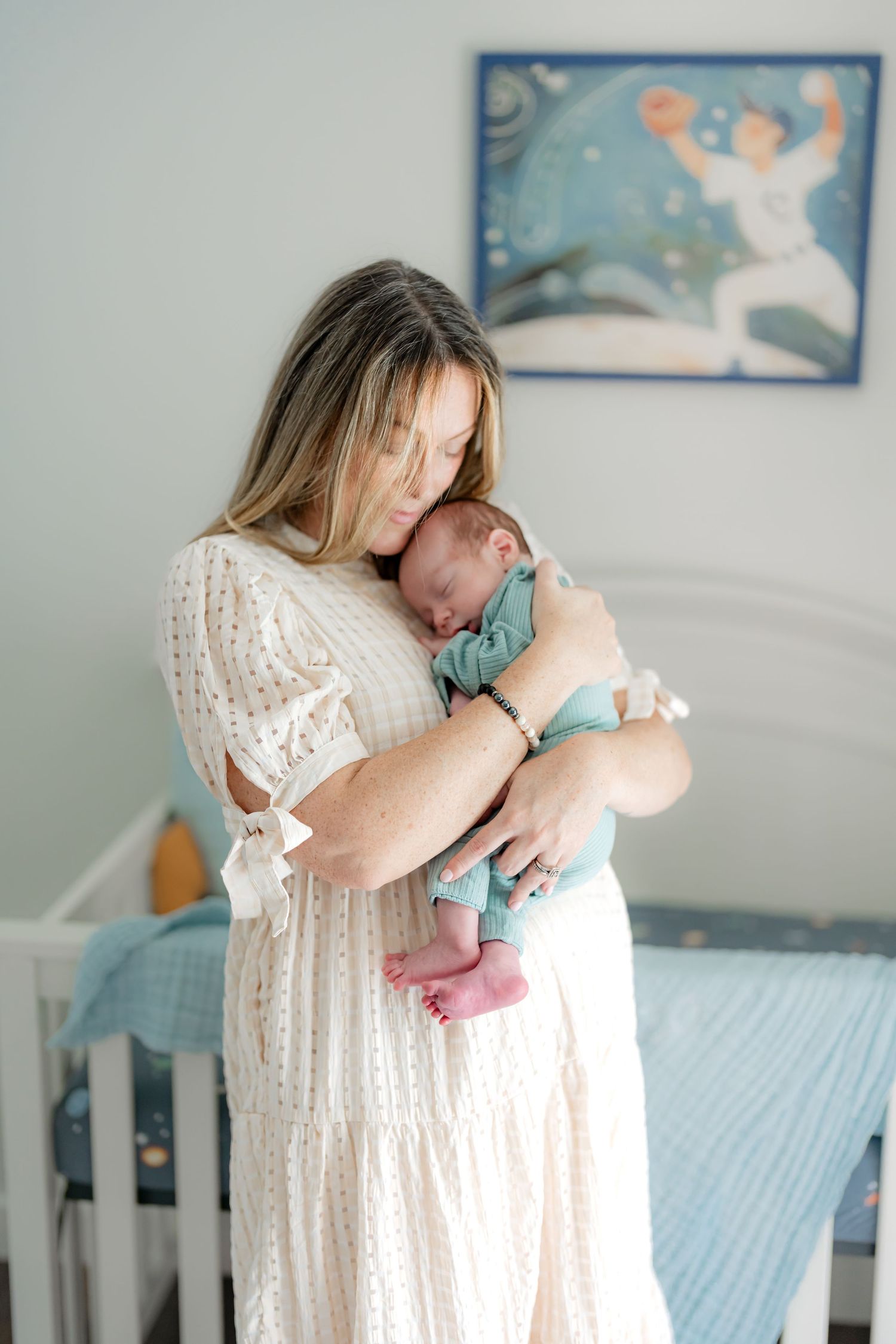 A woman in a white lace dress holds a sleeping baby in a nursery with blue decor.