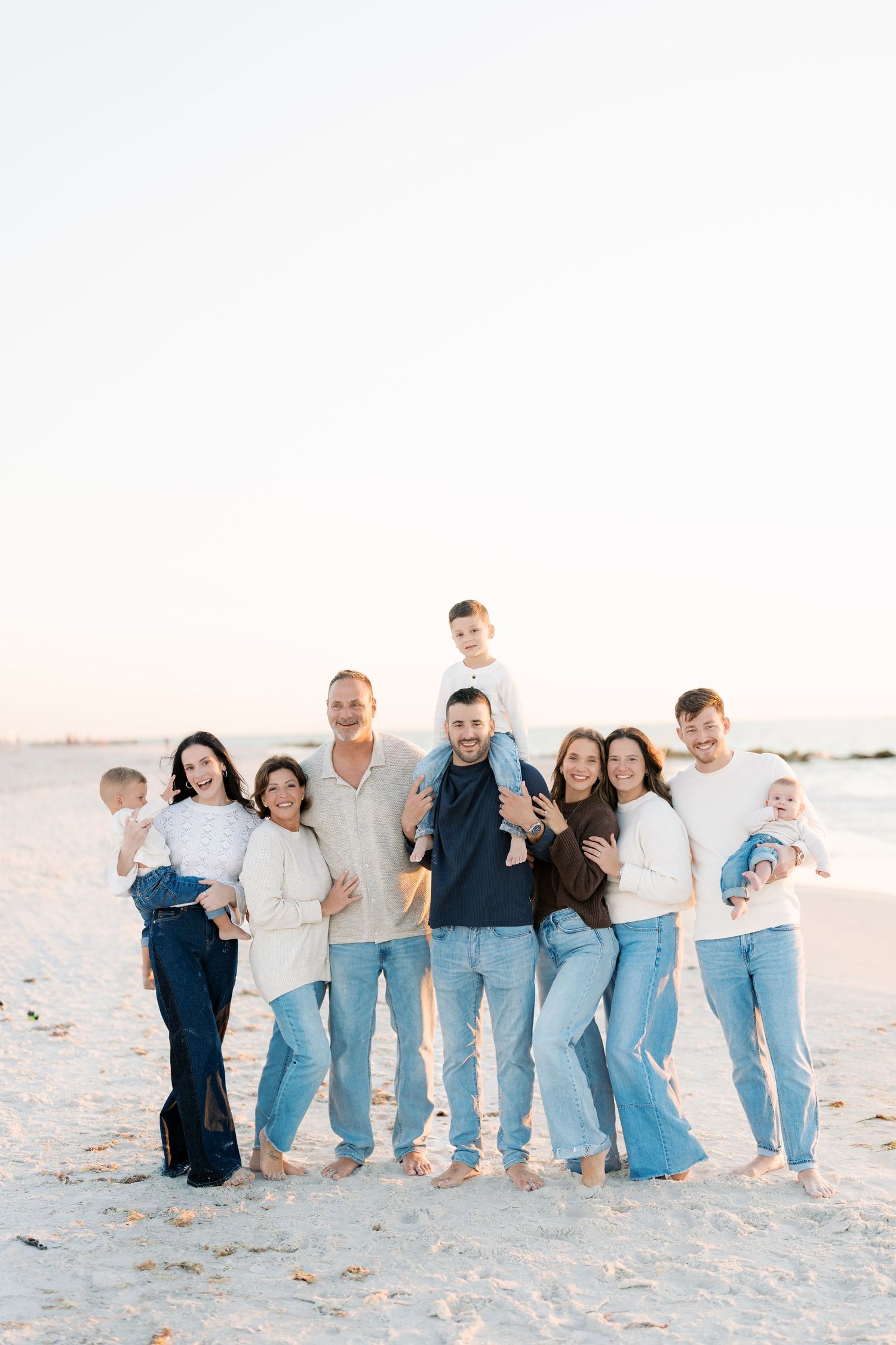 Large family group portrait on a sandy beach at sunset, wearing coordinated blue jeans and white and navy tops.