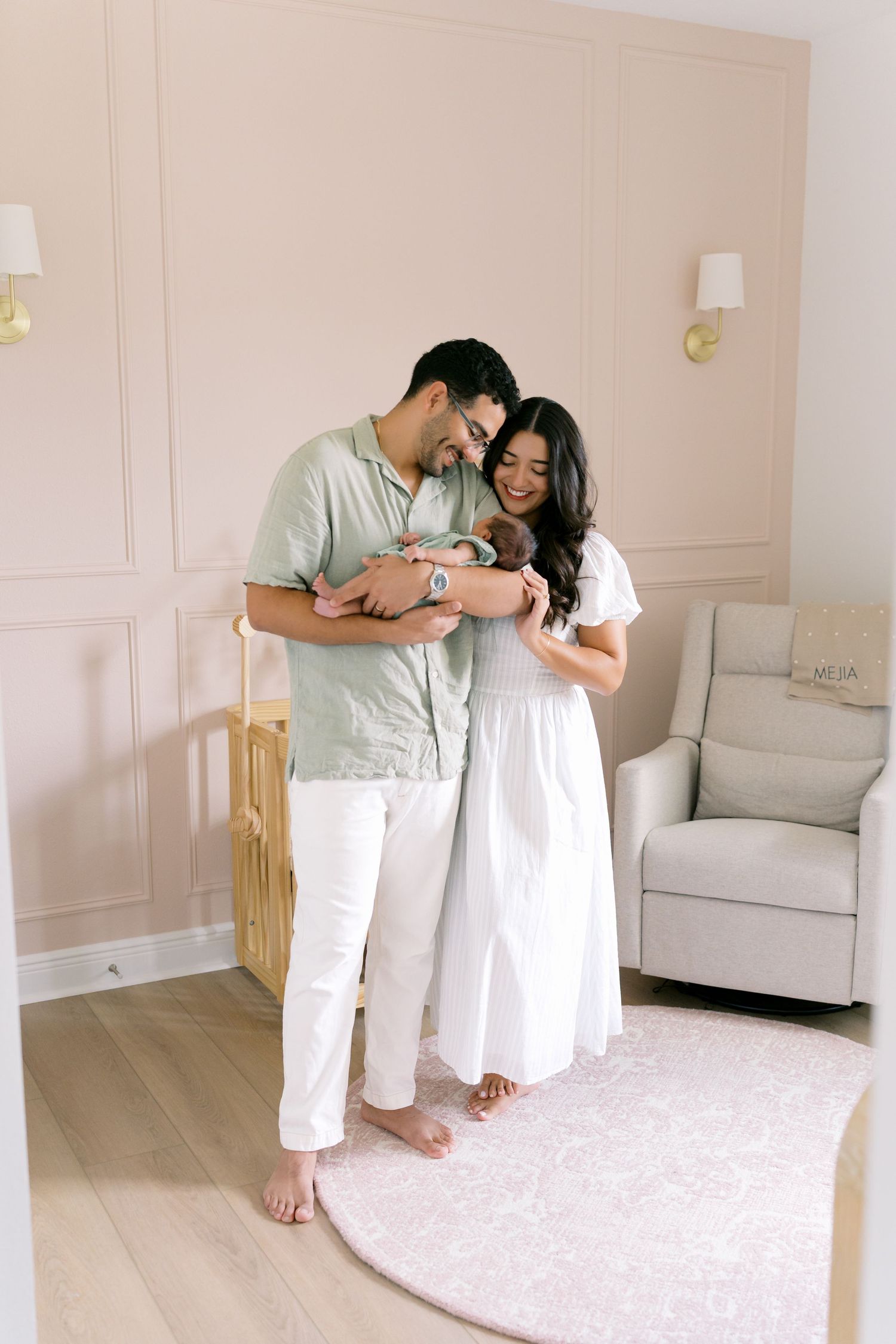 A couple in a pink nursery holding their newborn baby, standing beside a crib with a glider chair nearby.