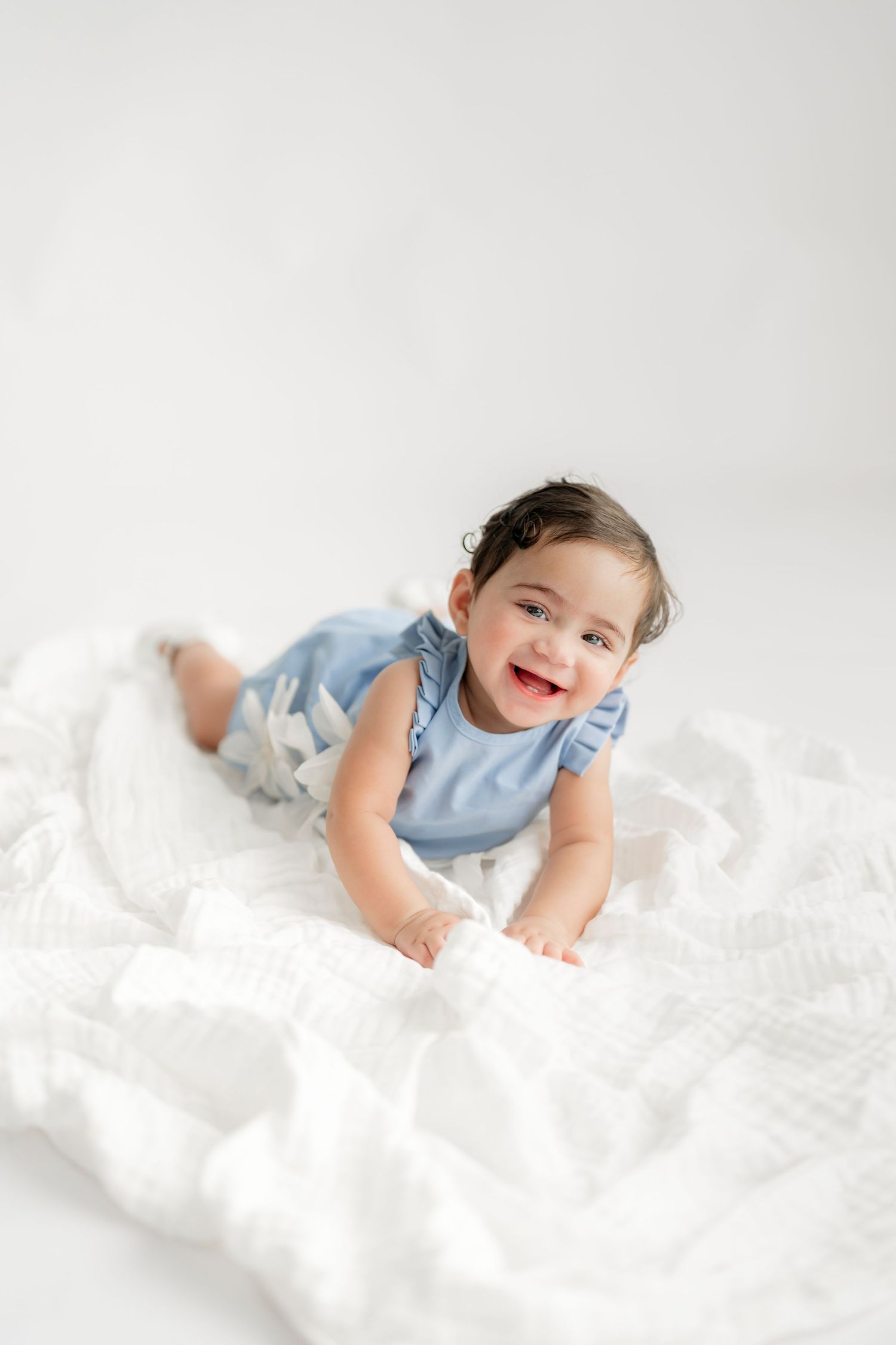 Baby in blue dress lying on white blanket smiling at camera.