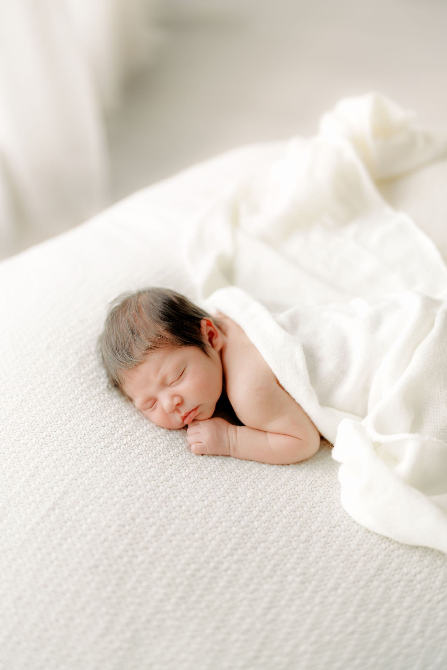 Peaceful newborn baby sleeping on white textured surface, wrapped in soft white blanket in bright airy room.