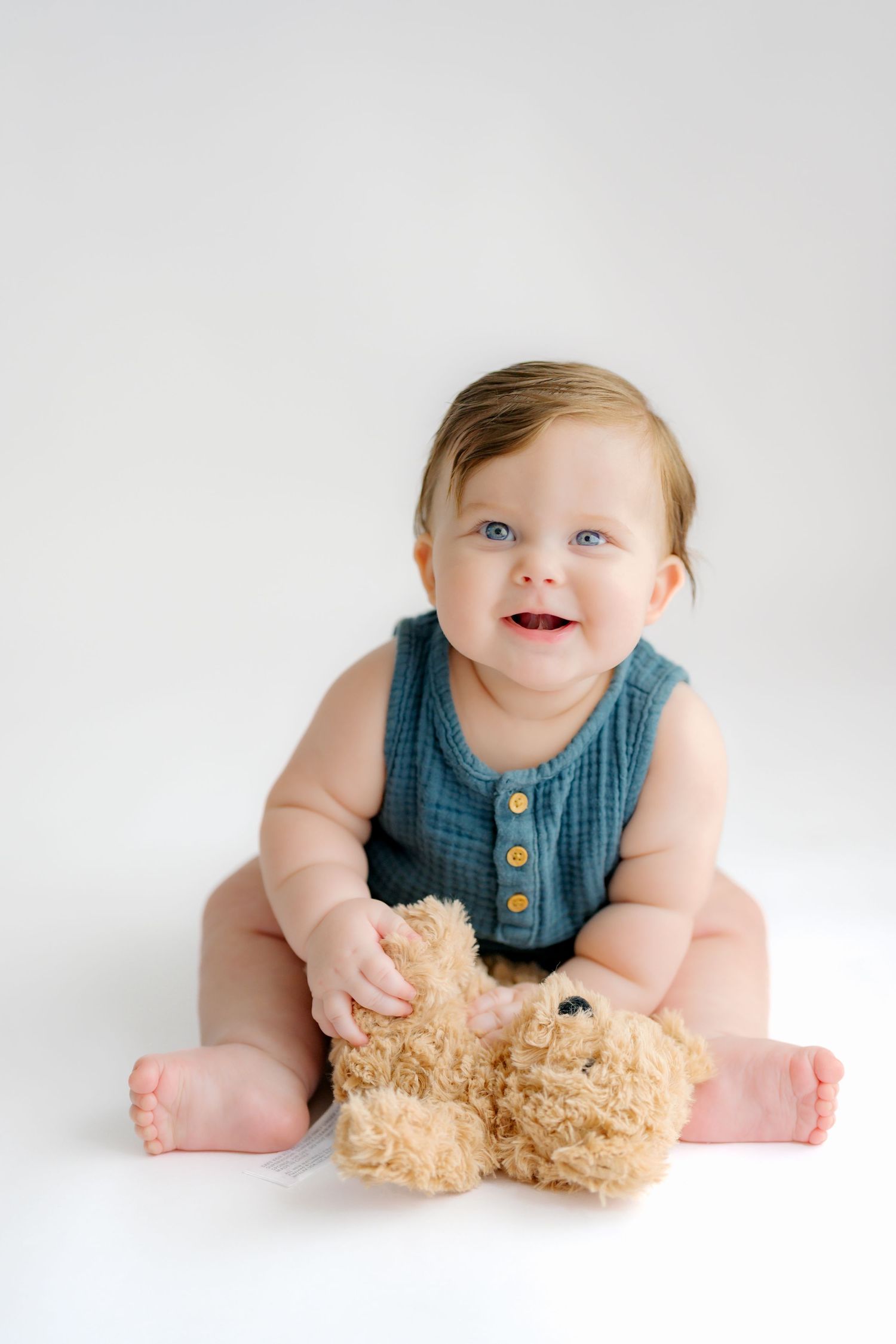 Baby in blue sleeveless outfit plays with soft beige teddy bear against white background in progressive photo sequence.