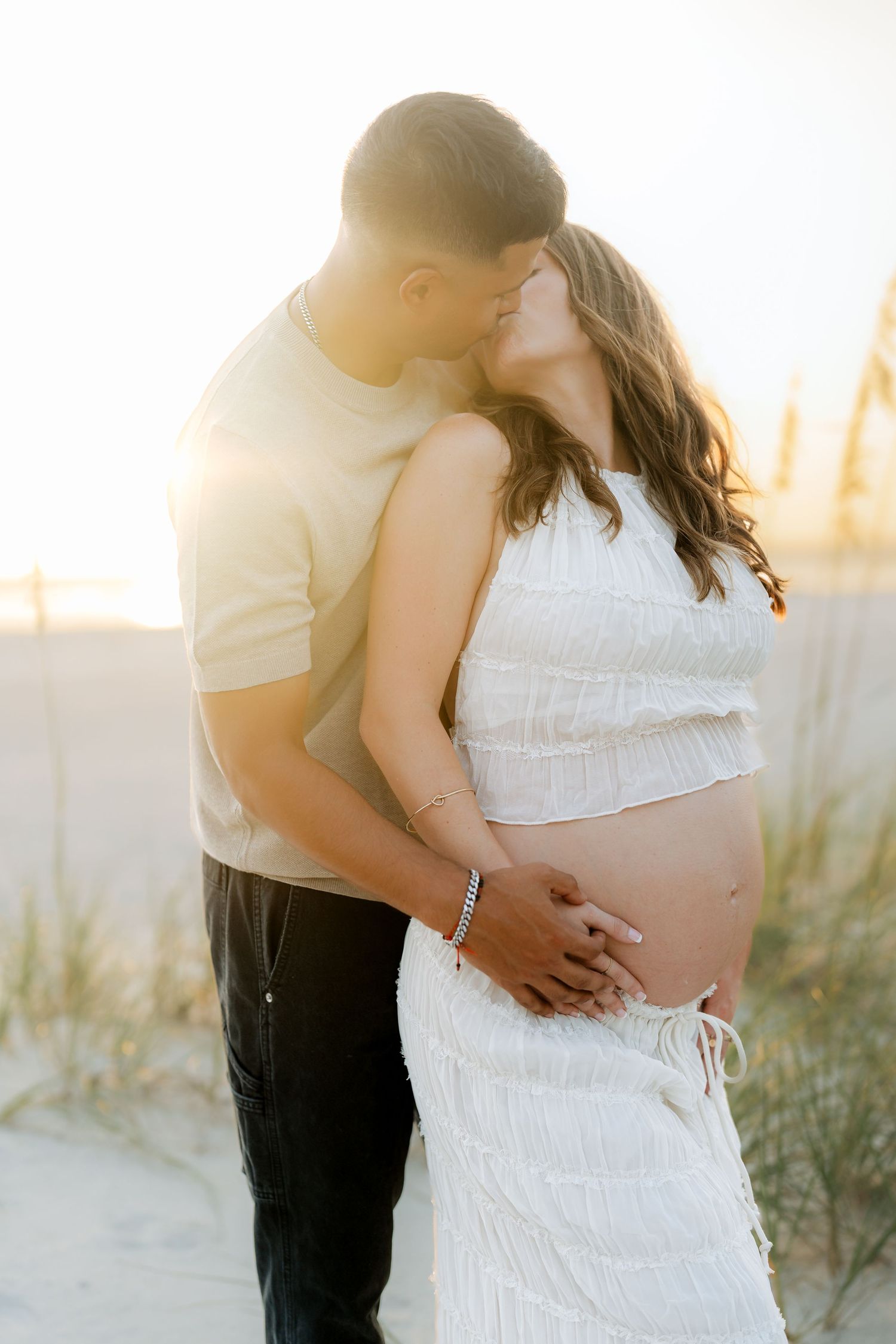 Couple embracing during sunset maternity shoot on the beach in white clothing.