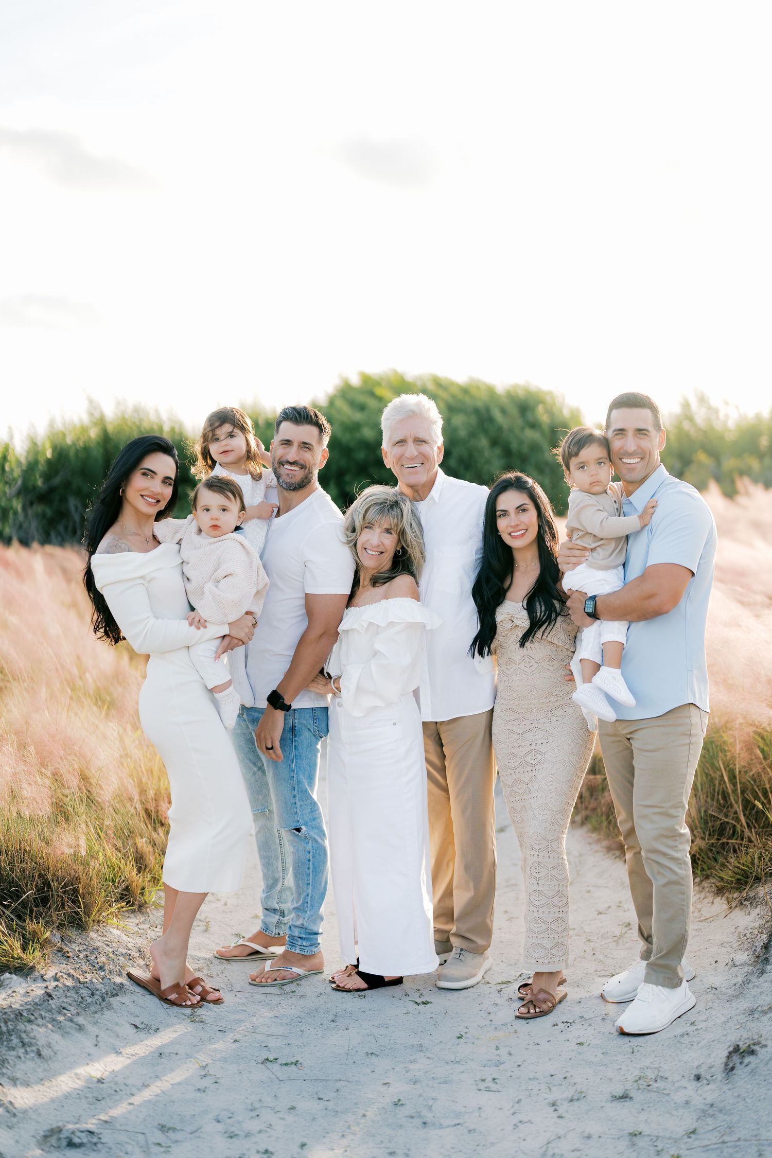 Extended family wearing white and neutral colors poses together on sandy beach at sunset.