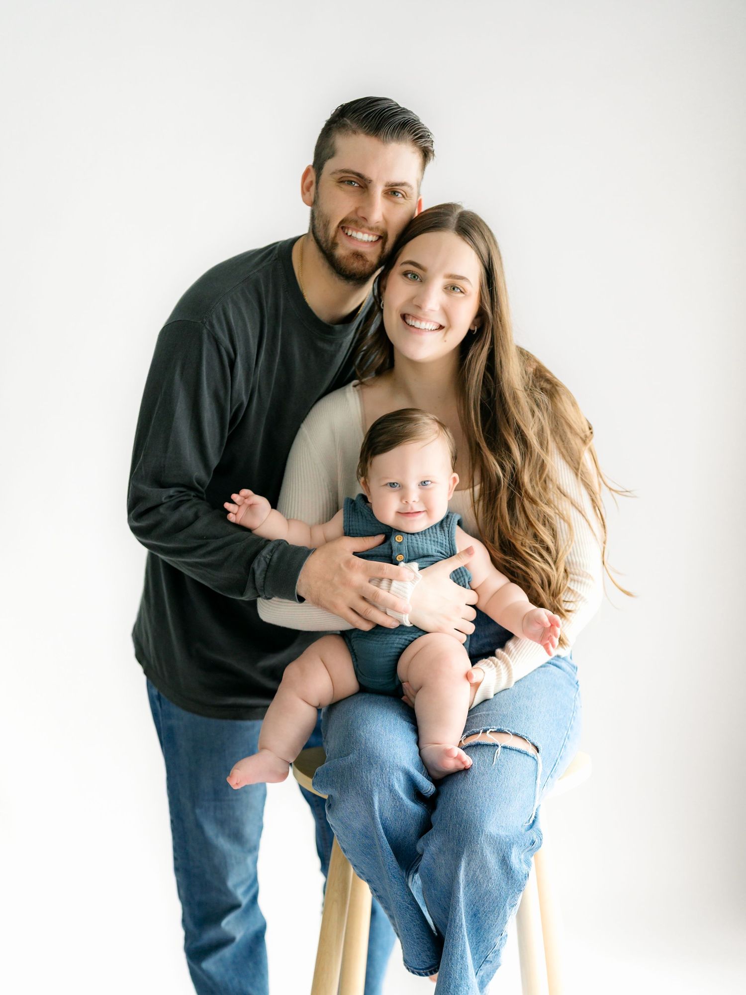 Family poses together against white background in casual attire with baby in blue outfit.