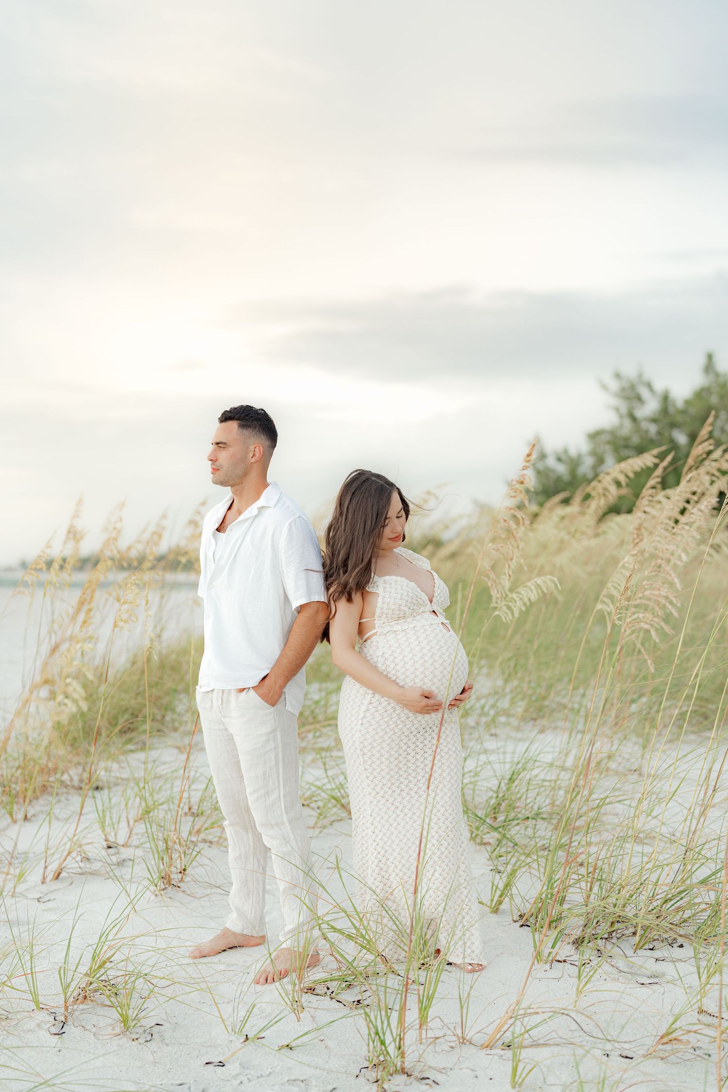 Beachside maternity photos with couple in white outfits among sea oats.