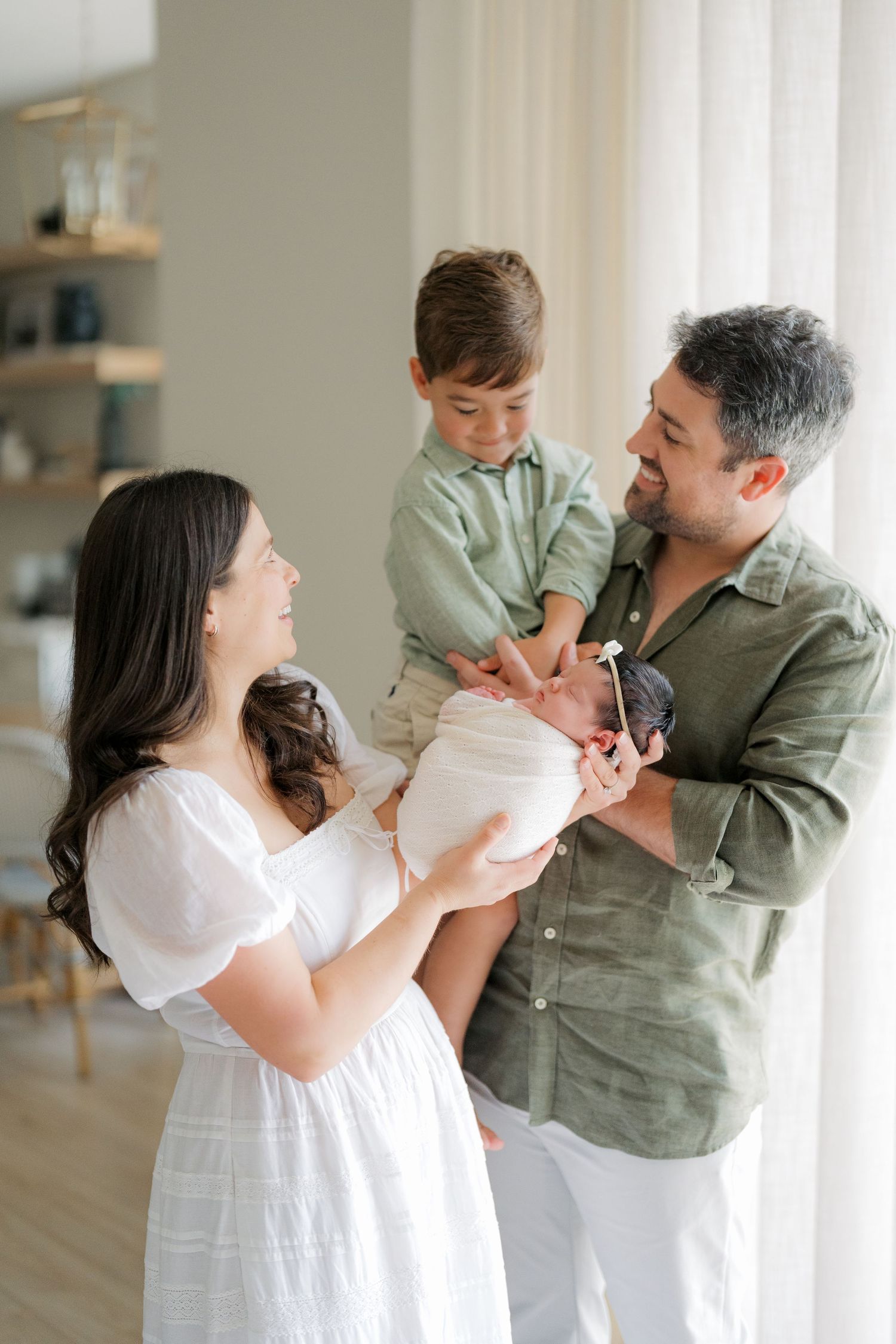 A happy family of four admiring their newborn baby, with mom in white dress and dad in green shirt holding toddler son.