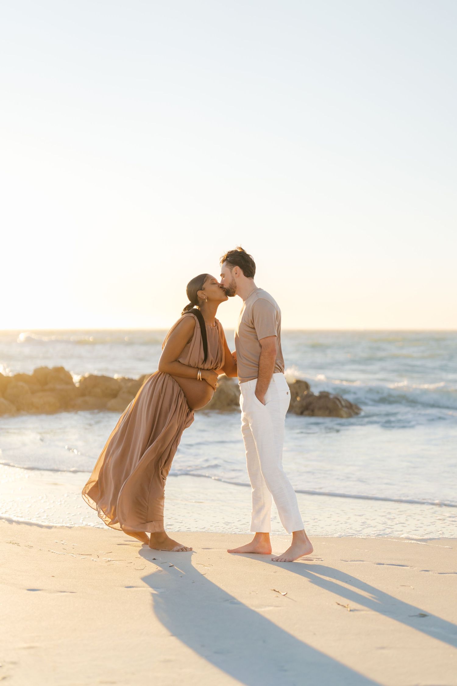 Interracial couple sharing a kiss on sandy beach during golden sunset by rocky shoreline.
