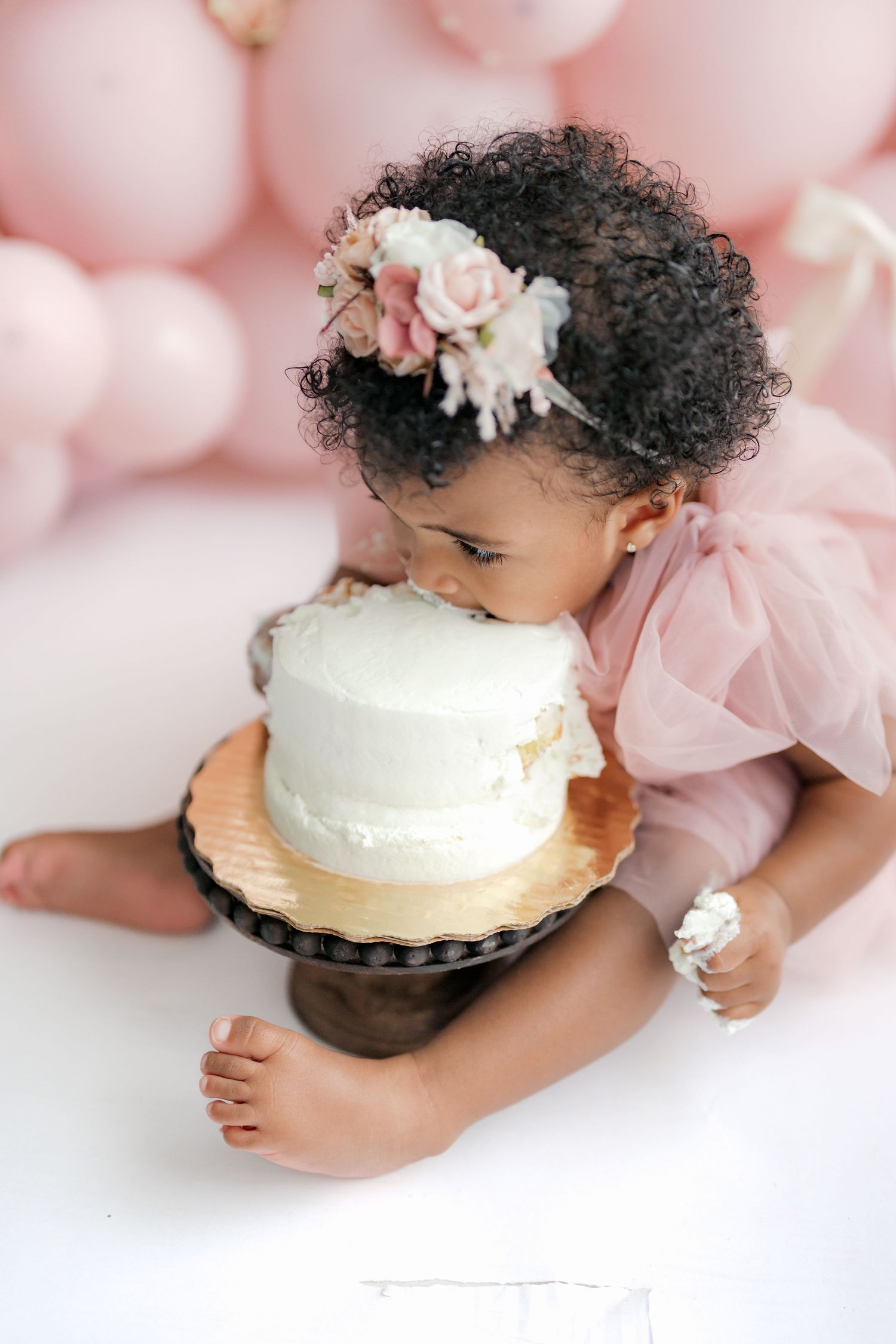 Baby in pink dress reaches for white frosted cake during first birthday cake smash photoshoot.