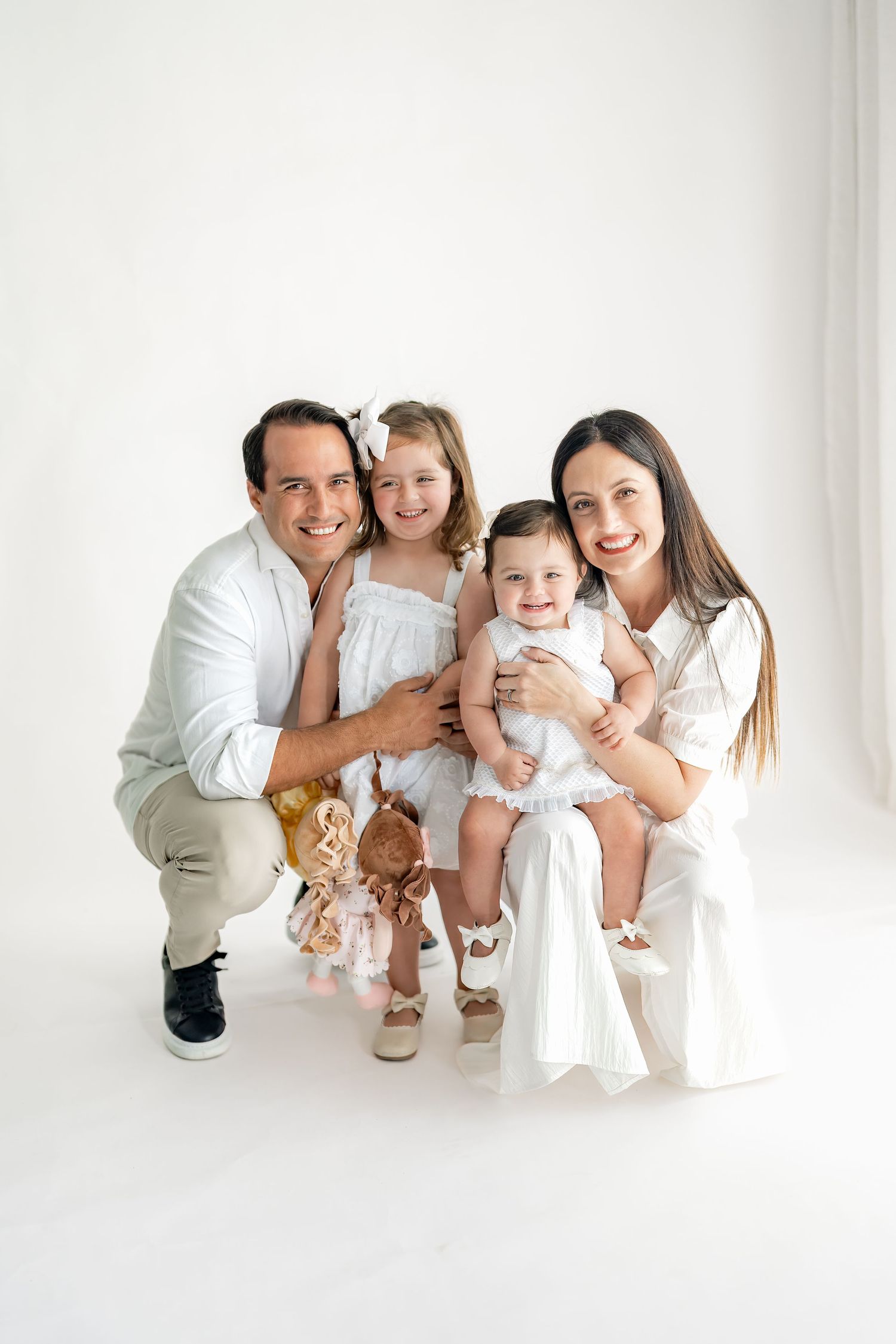 Family wearing white outfits crouches together against white wall in minimalist portrait session.