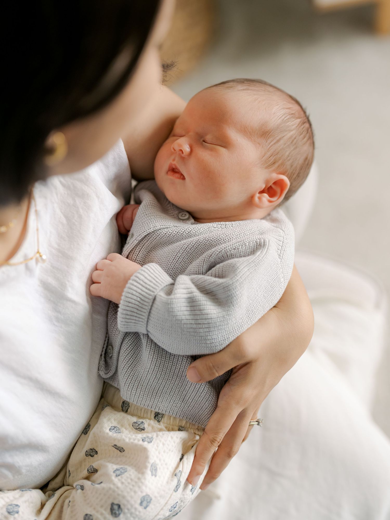 A mother tenderly holds her sleeping newborn baby dressed in a soft gray knit cardigan and floral pants.