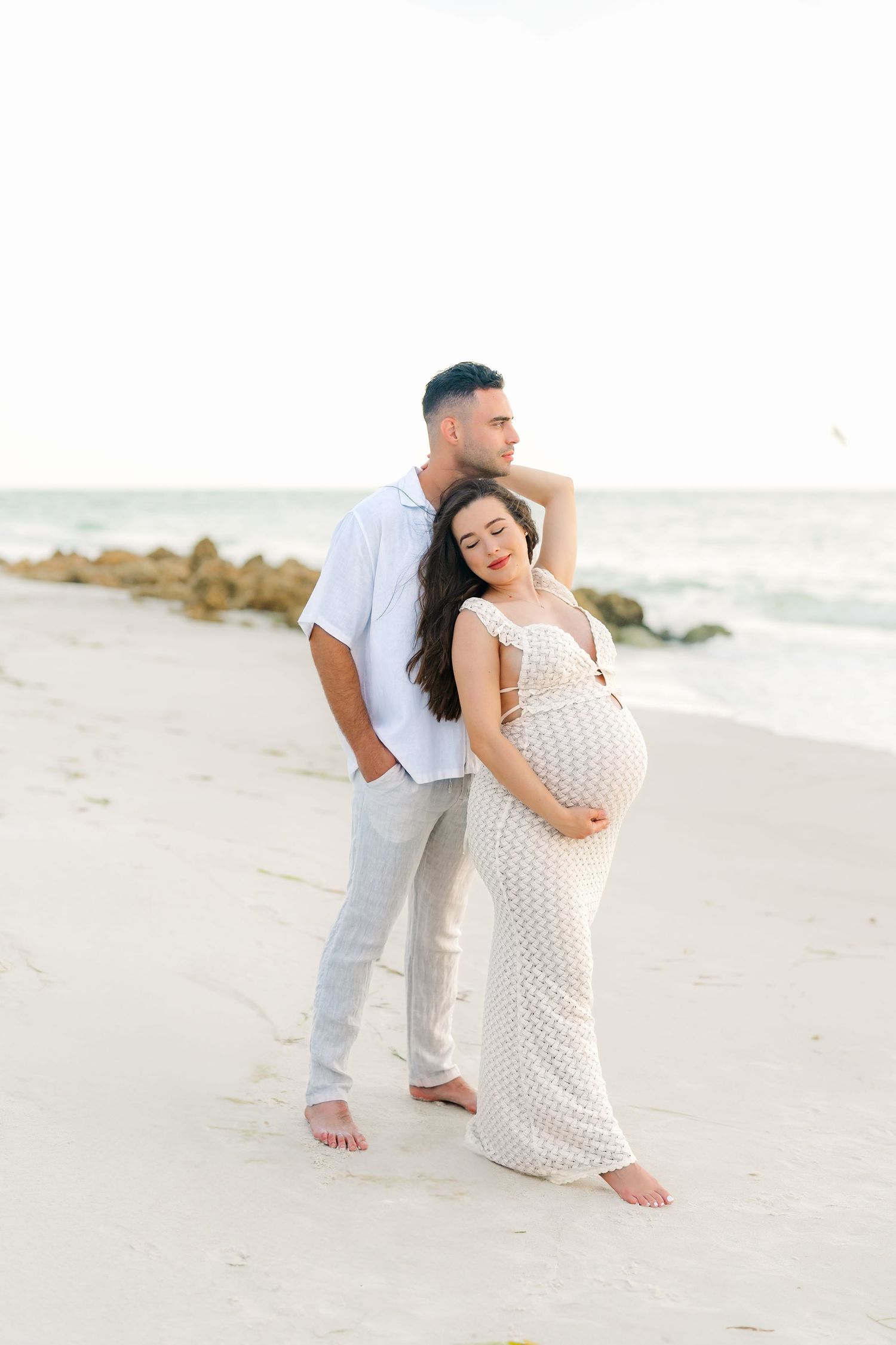 Maternity beach photo session with couple in white outfits posing on sandy shore.