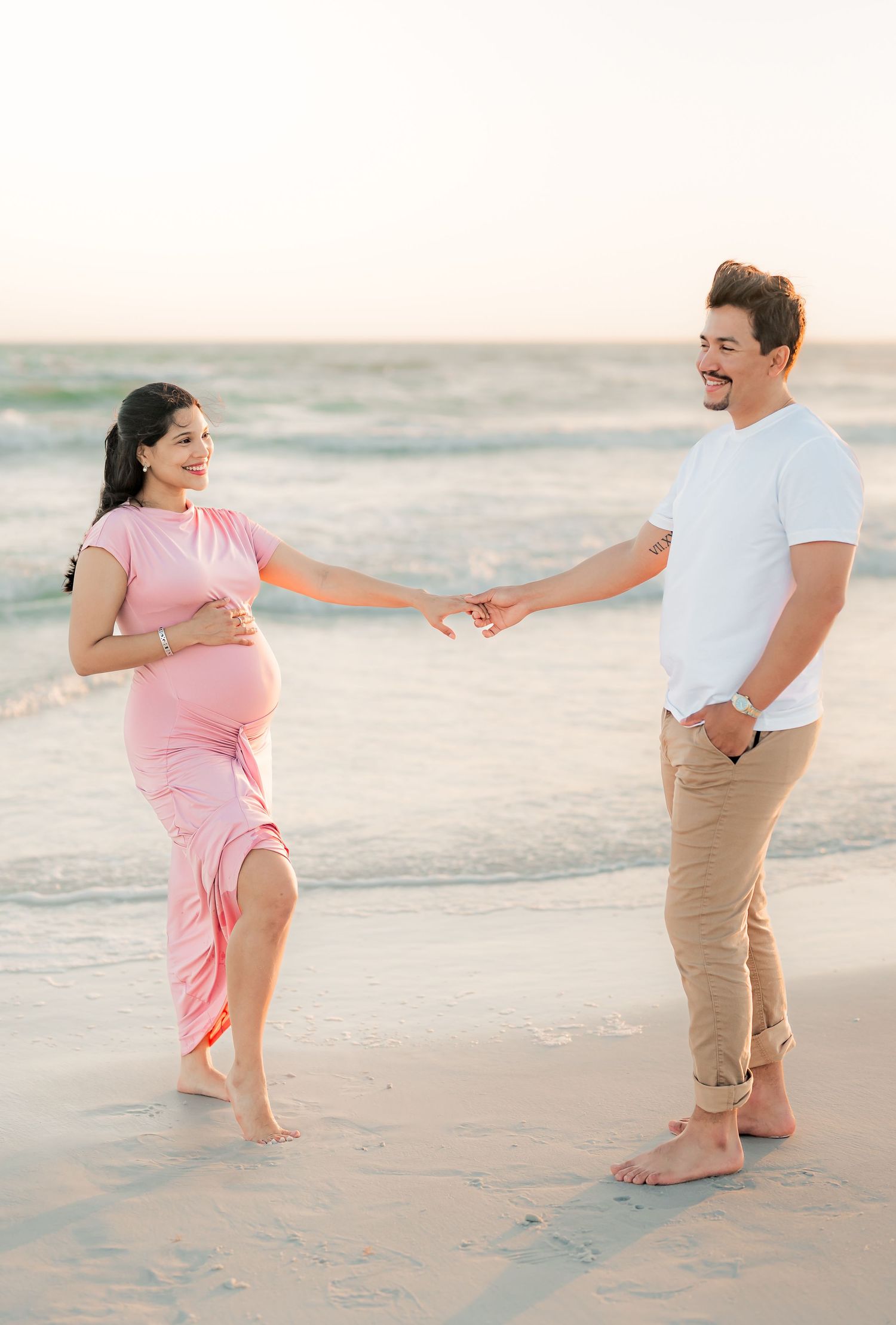 A couple in pink and white outfits dances playfully together on a sandy beach at sunset.