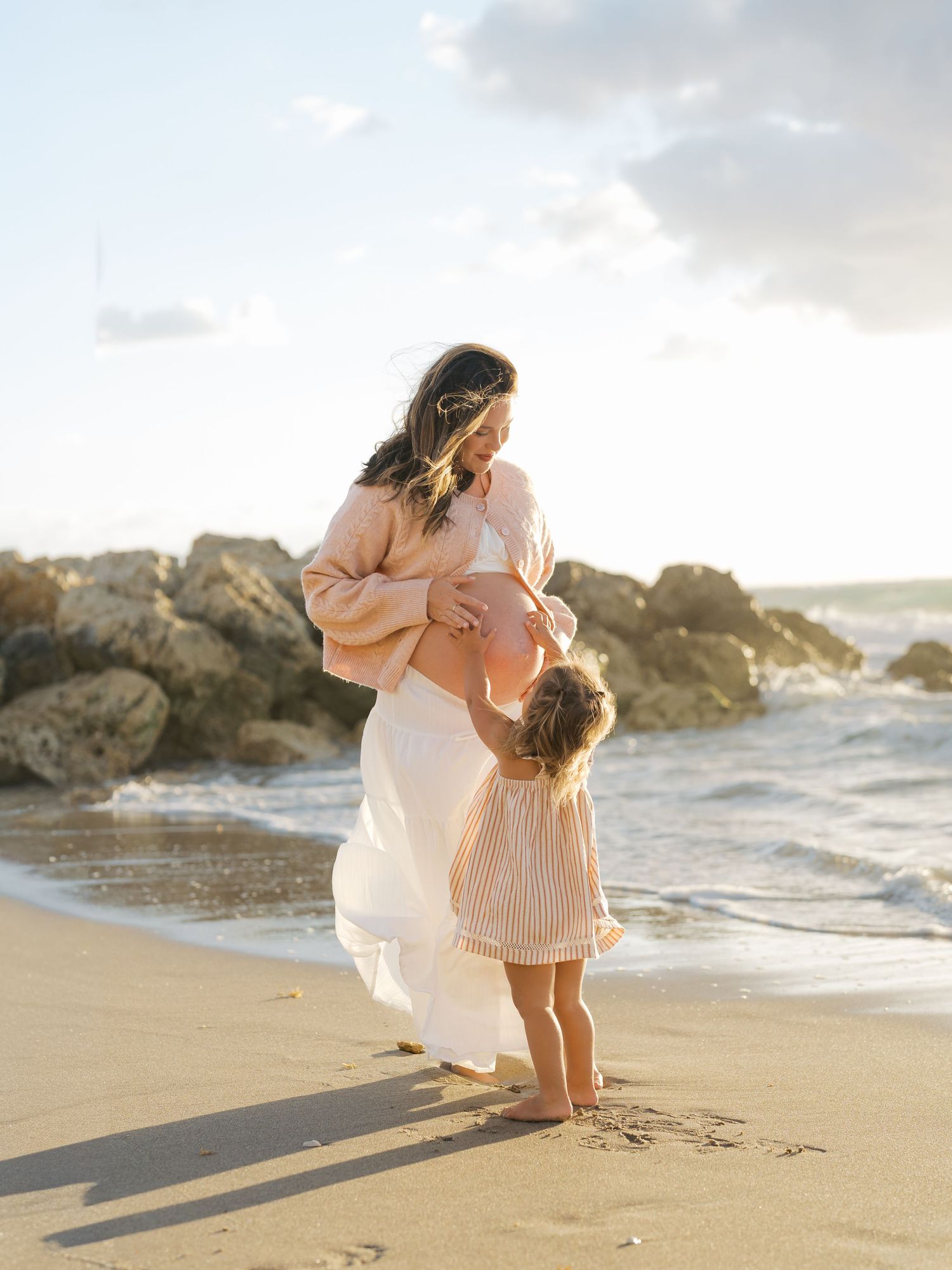 Pregnant woman in pink top and white skirt with toddler on sunny beach near rocky shoreline.