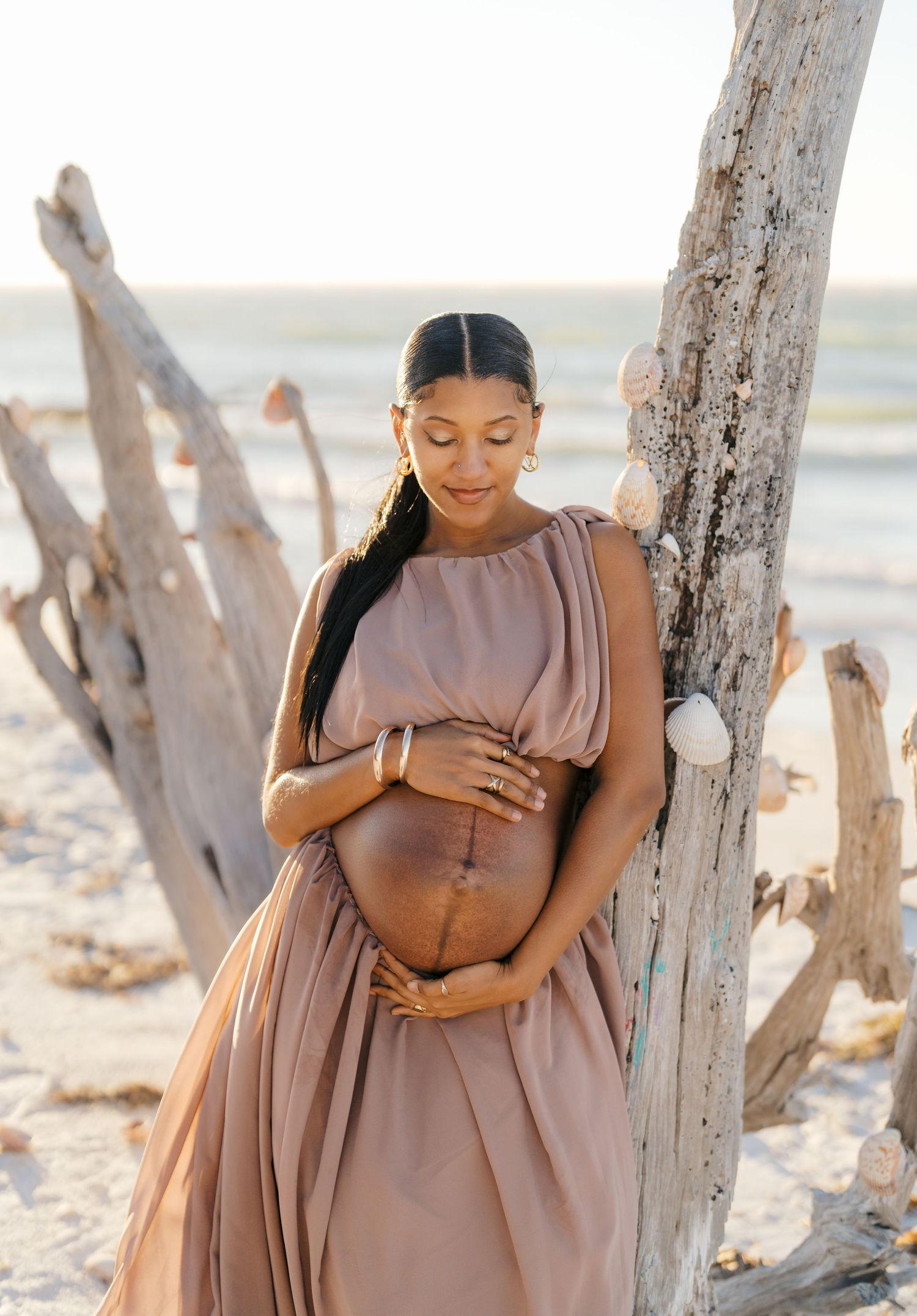 Pregnant woman in a flowing mauve dress cradles her belly on a beach, leaning against driftwood at golden hour.