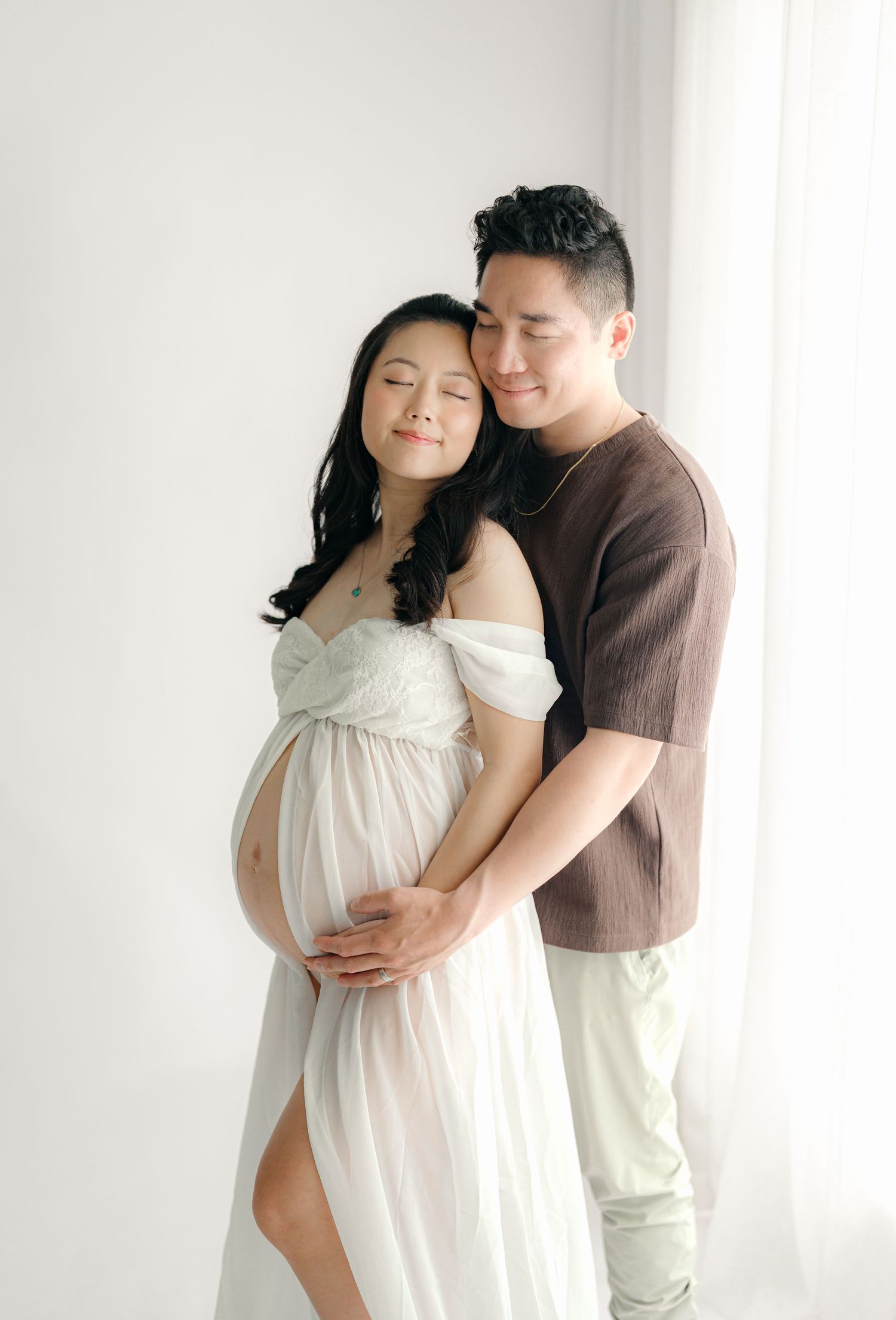 Indoor maternity photoshoot with couple in white dress and brown shirt against bright background.