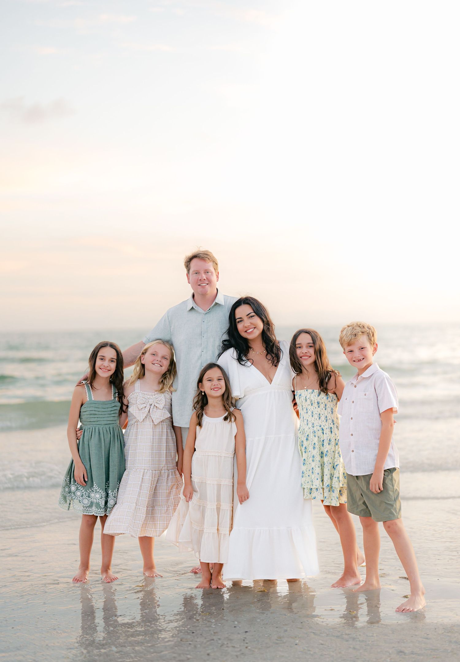 Family group portrait on beach at sunset wearing coordinated white and pastel outfits.