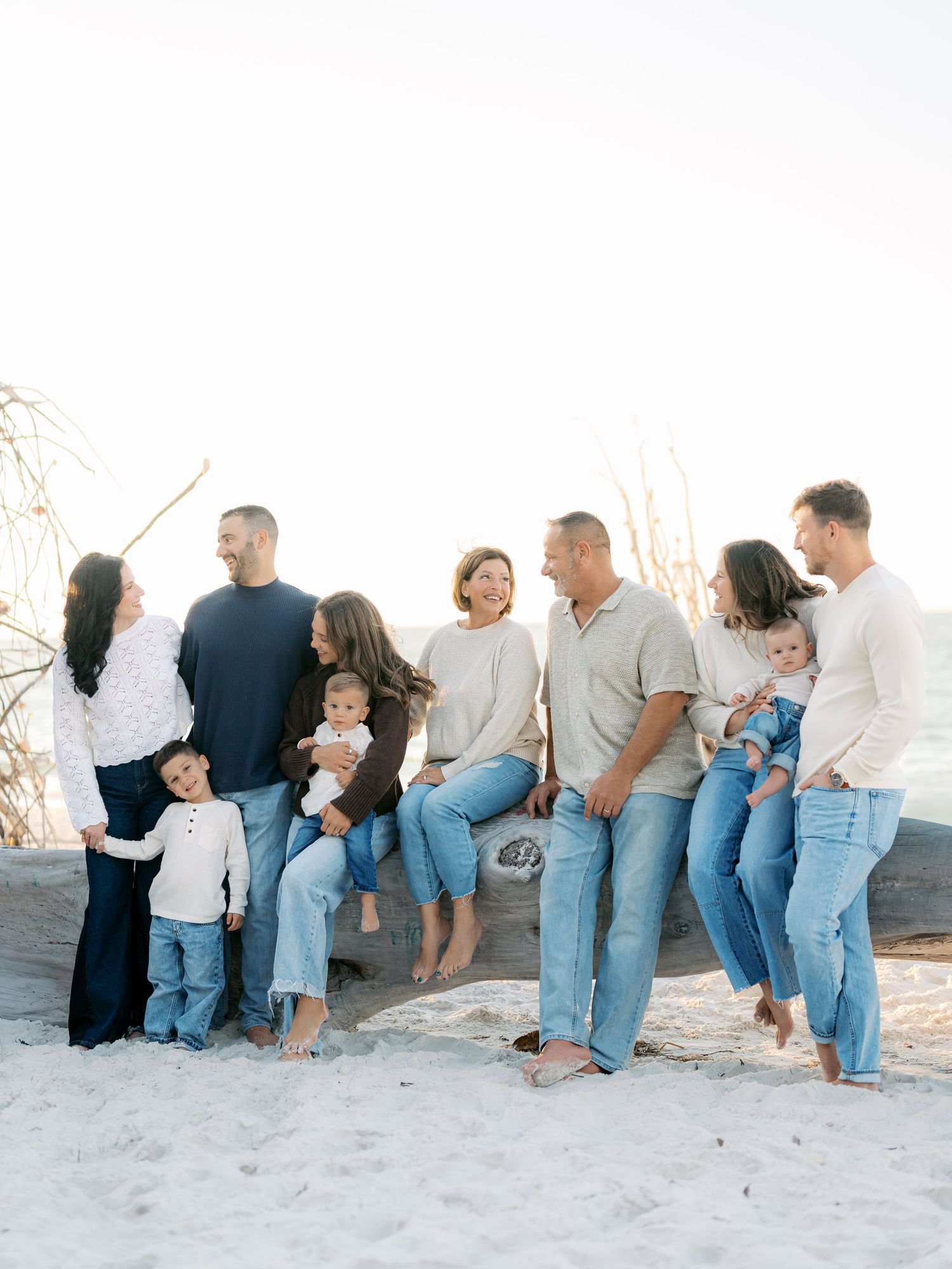 A group of people sitting casually on a white wall outdoors wearing jeans and neutral colored tops.