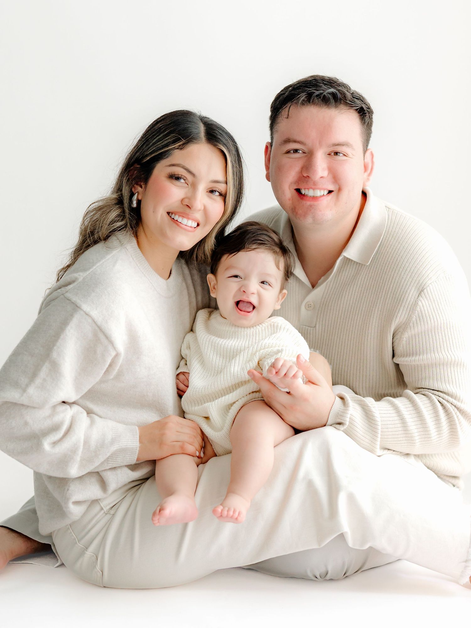 A family wearing coordinated cream and white outfits poses together on the floor in a minimalist studio setting.