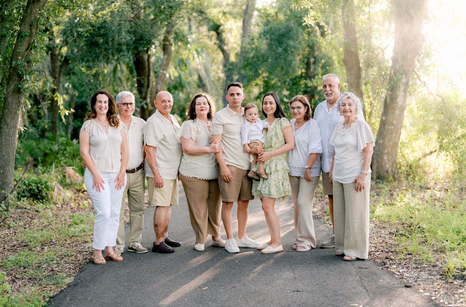 Group of people in coordinated white and beige outfits pose together for outdoor family photos in a sunlit wooded area.