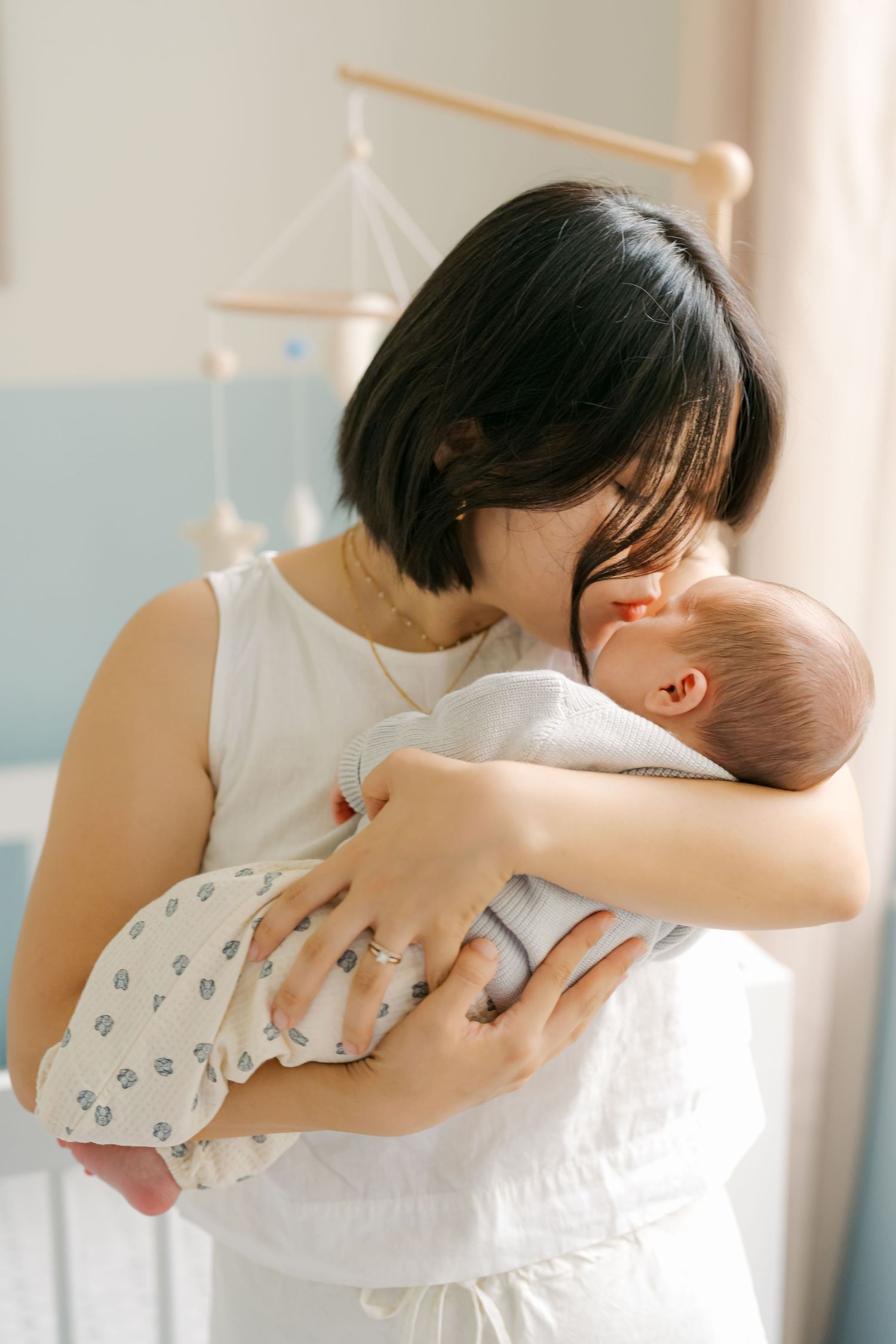 A mother with short dark hair tenderly holds and kisses her newborn baby in a softly lit nursery room.