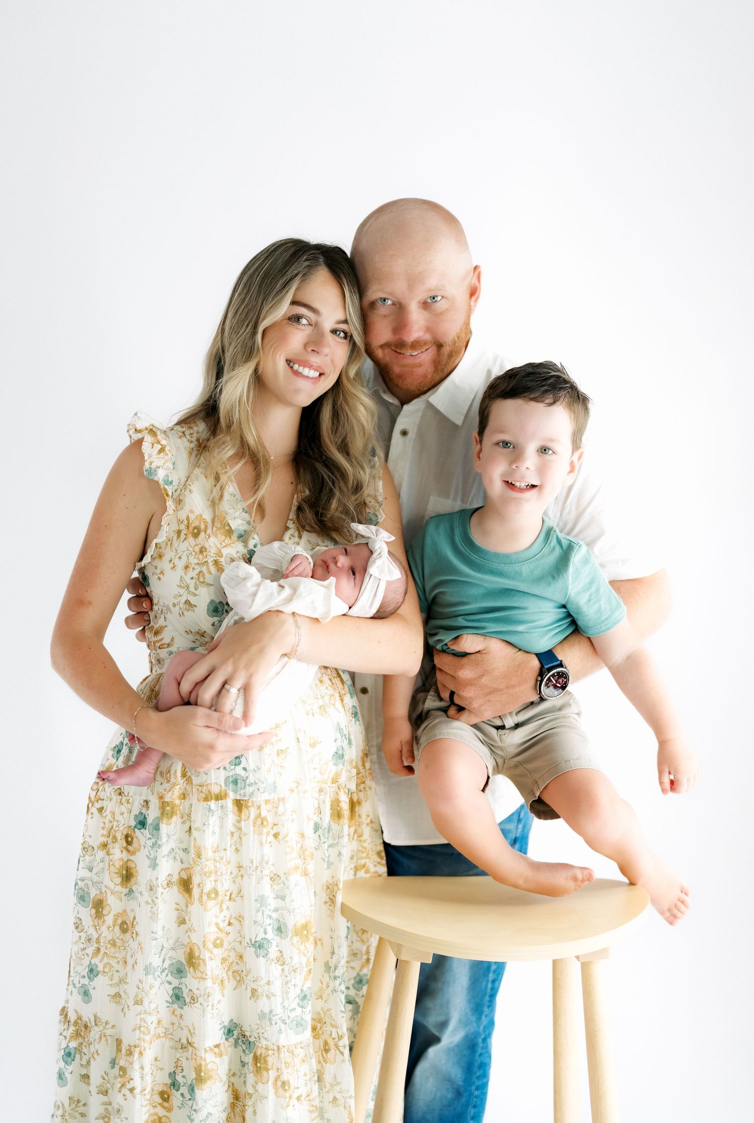 Happy family of four posing in studio, mother holding newborn, father lifting toddler son, white background.
