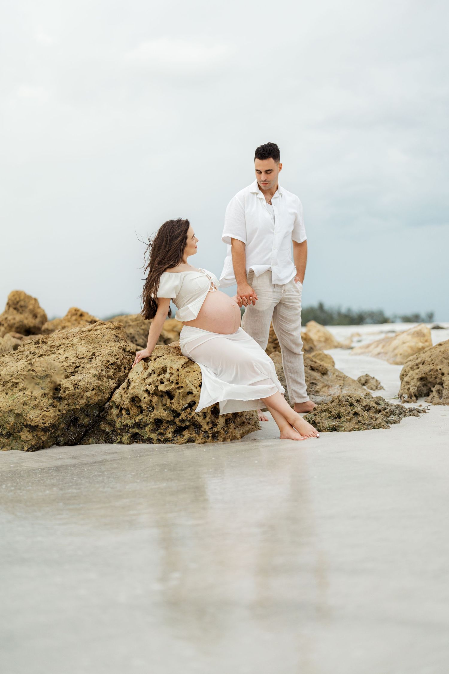 Couple in white attire sitting together on coastal rocks during a beach maternity photoshoot.