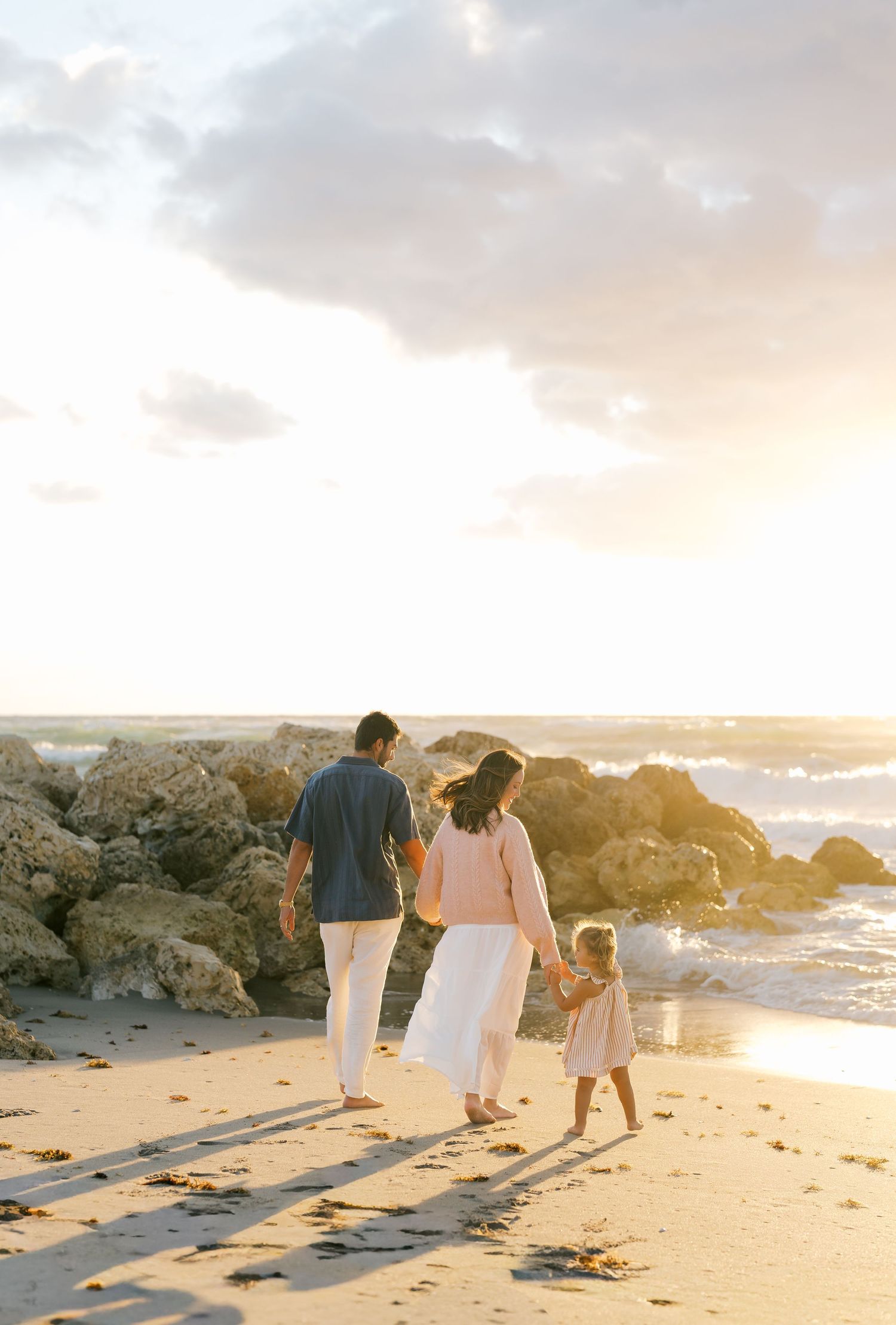 Family of three walking hand-in-hand on a sandy beach at golden sunset with rocky shore and ocean waves.