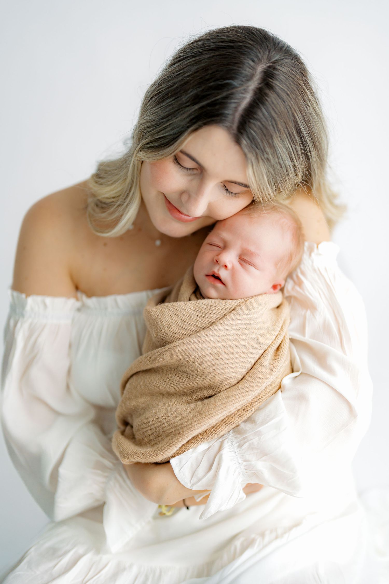 Mother in white dress tenderly holds sleeping newborn wrapped in beige blanket against white background.