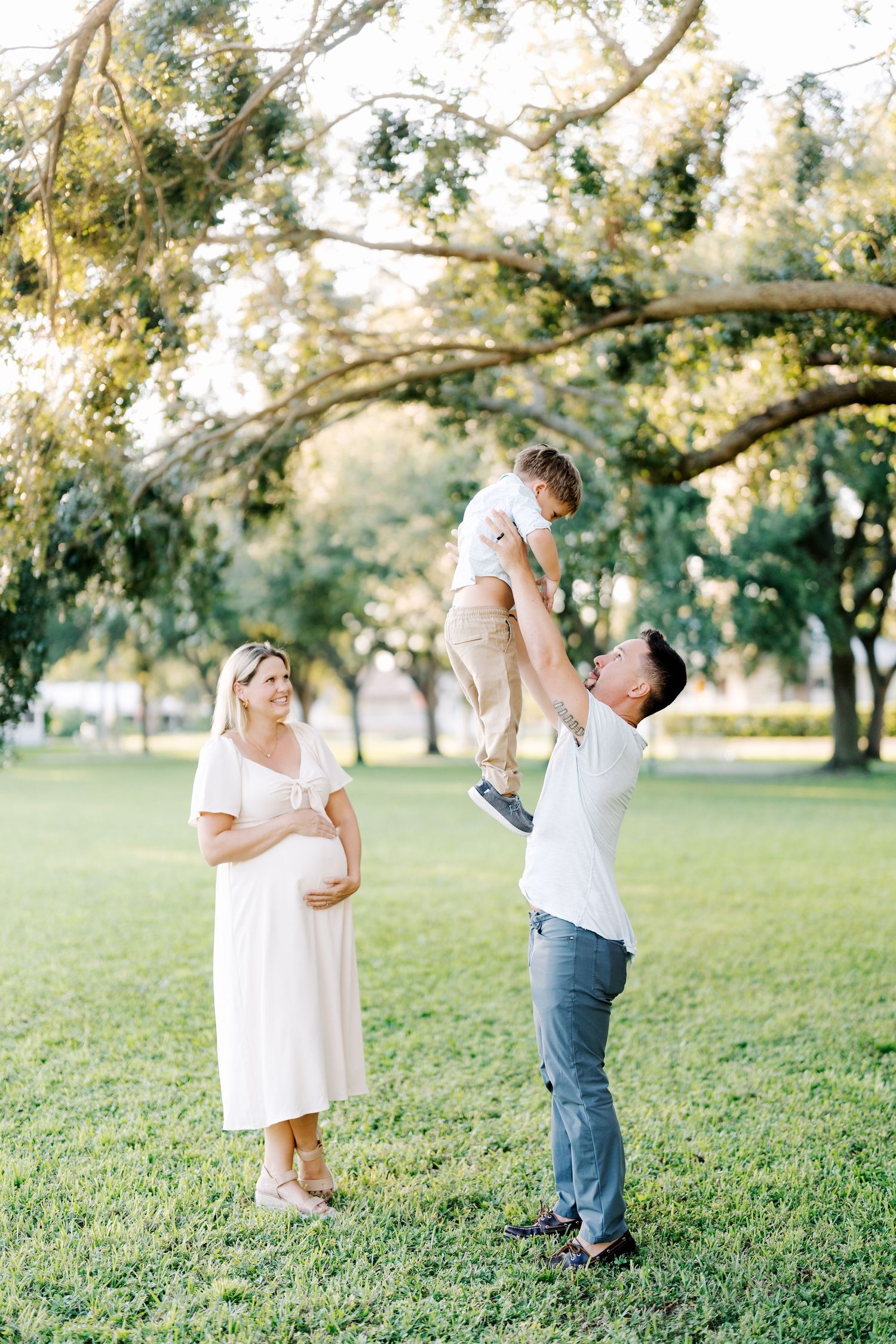 Pregnant woman in white dress watches husband toss toddler in air during outdoor maternity photo session in green park.