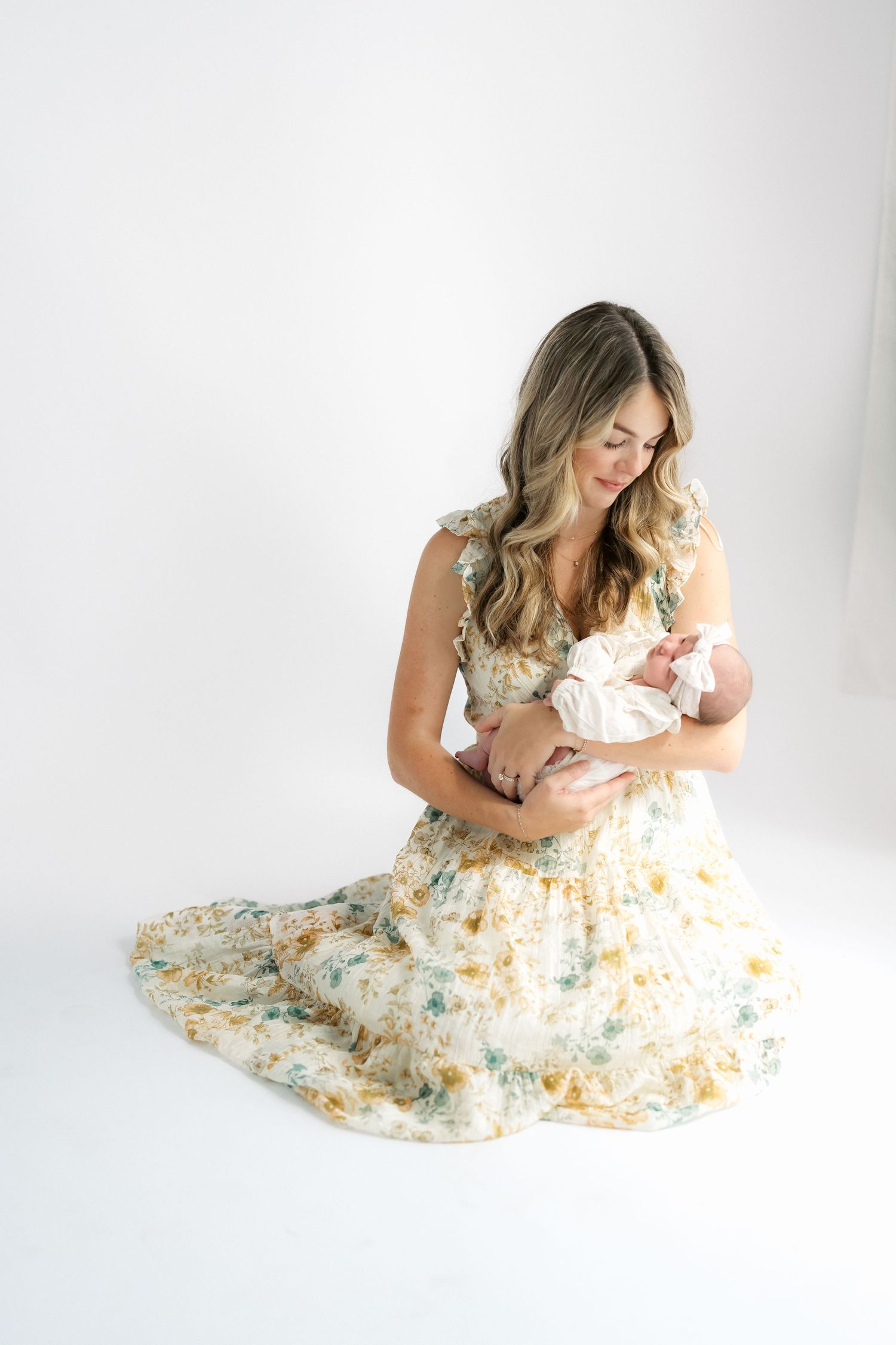 Woman in floral dress sitting on white background, lovingly cradling a newborn baby in her arms.