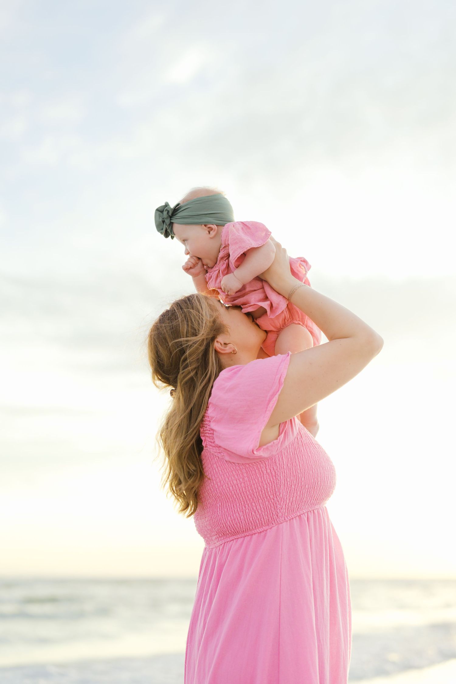 Mother in pink dress lifts baby in matching outfit during beach sunset photoshoot.