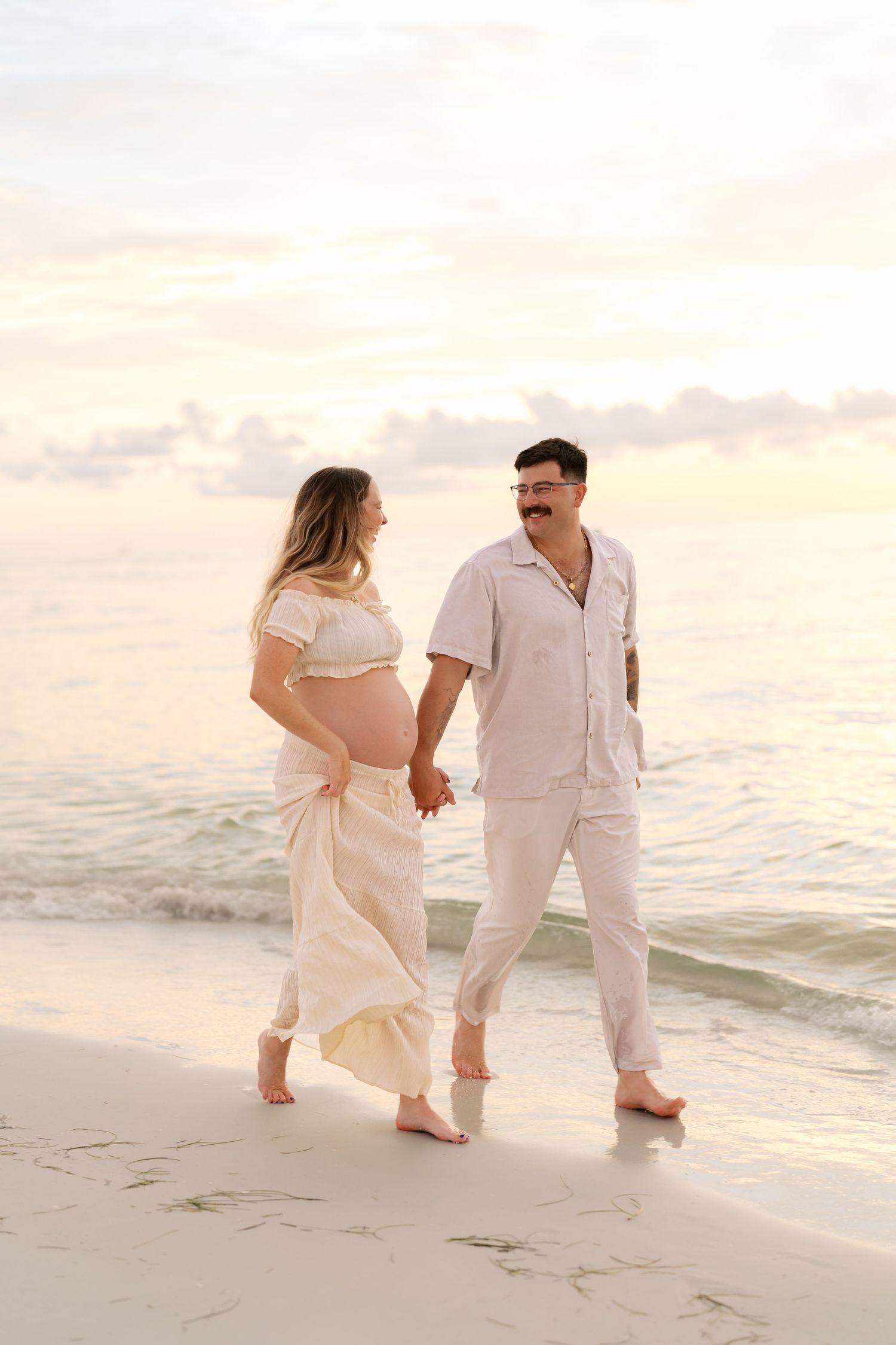 Pregnant couple holding hands and walking barefoot on a sandy beach at sunset in matching cream outfits.