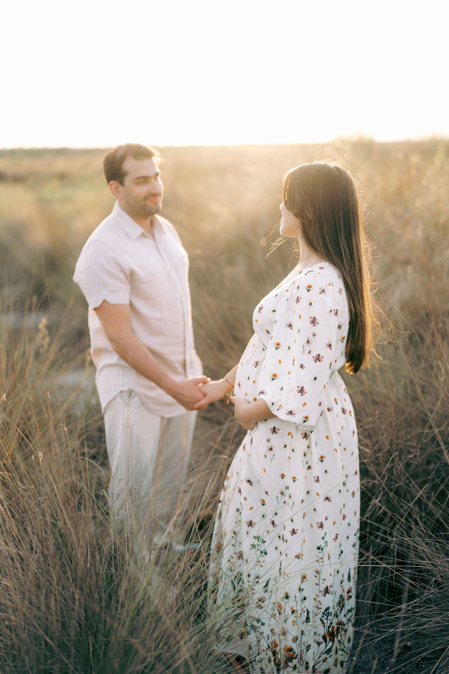 Sunset maternity session in tall grass with couple wearing white and floral outfits.