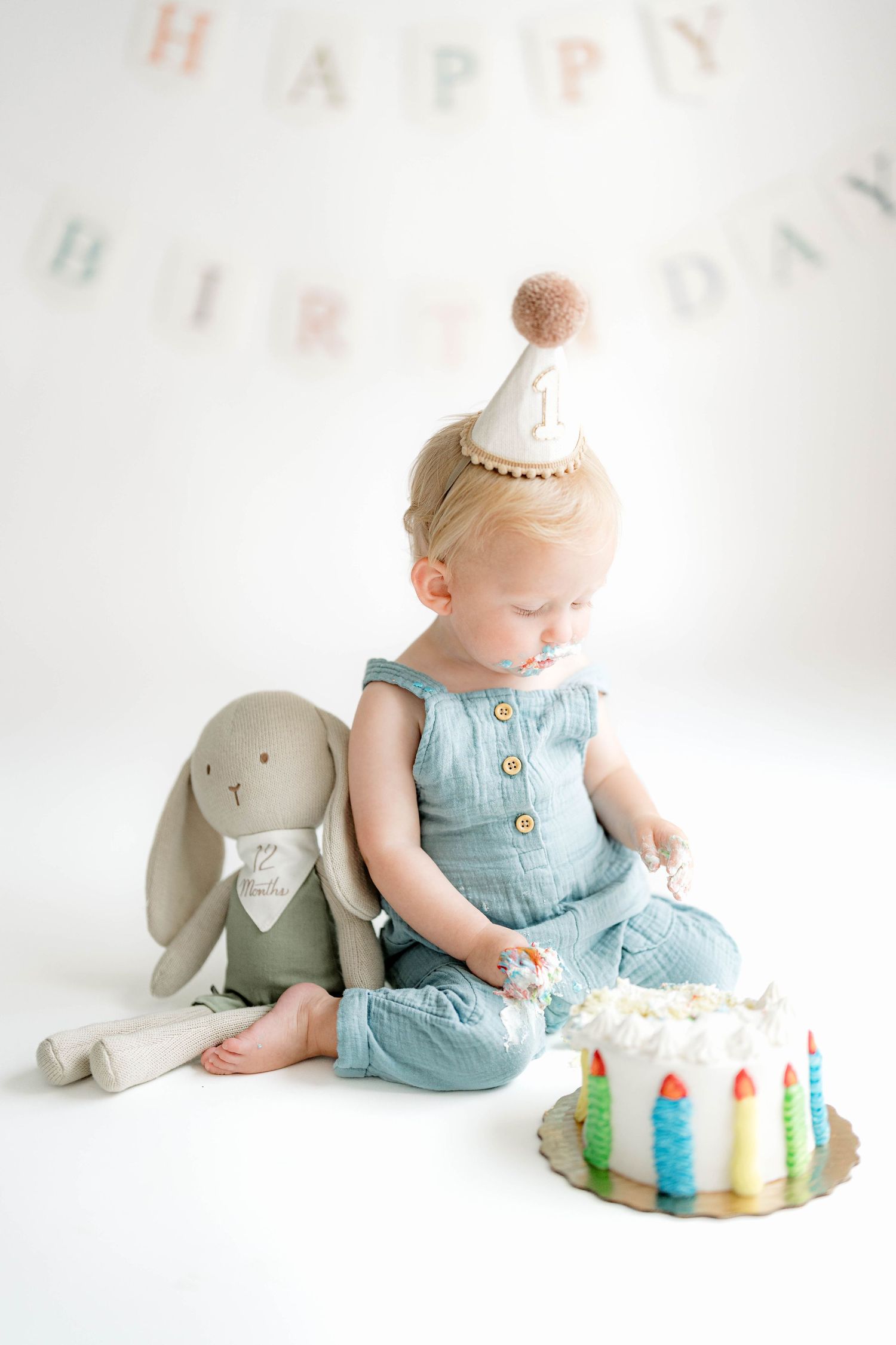 Baby in light blue romper sits beside birthday cake and stuffed elephant toy in minimalist white studio setting.