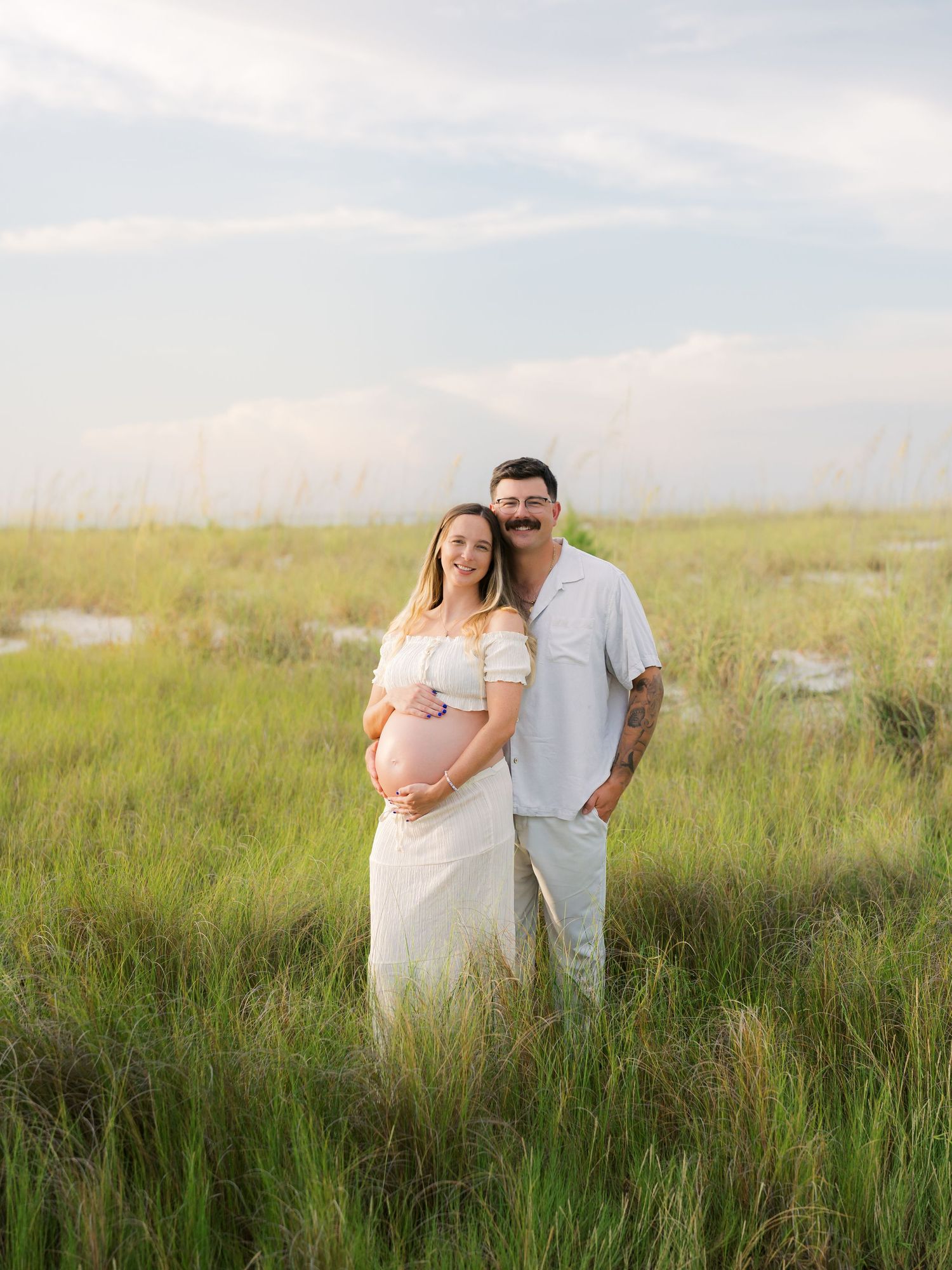 Pregnant couple in white outfits posing in a lush green field during golden hour for a maternity photo session.